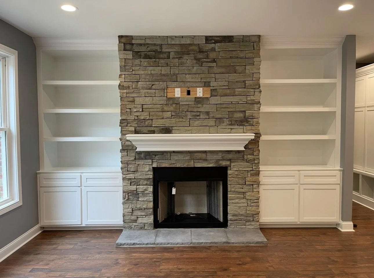 Living room with a stone fireplace, white built-in shelves on either side, a wooden floor, and a window on the left.