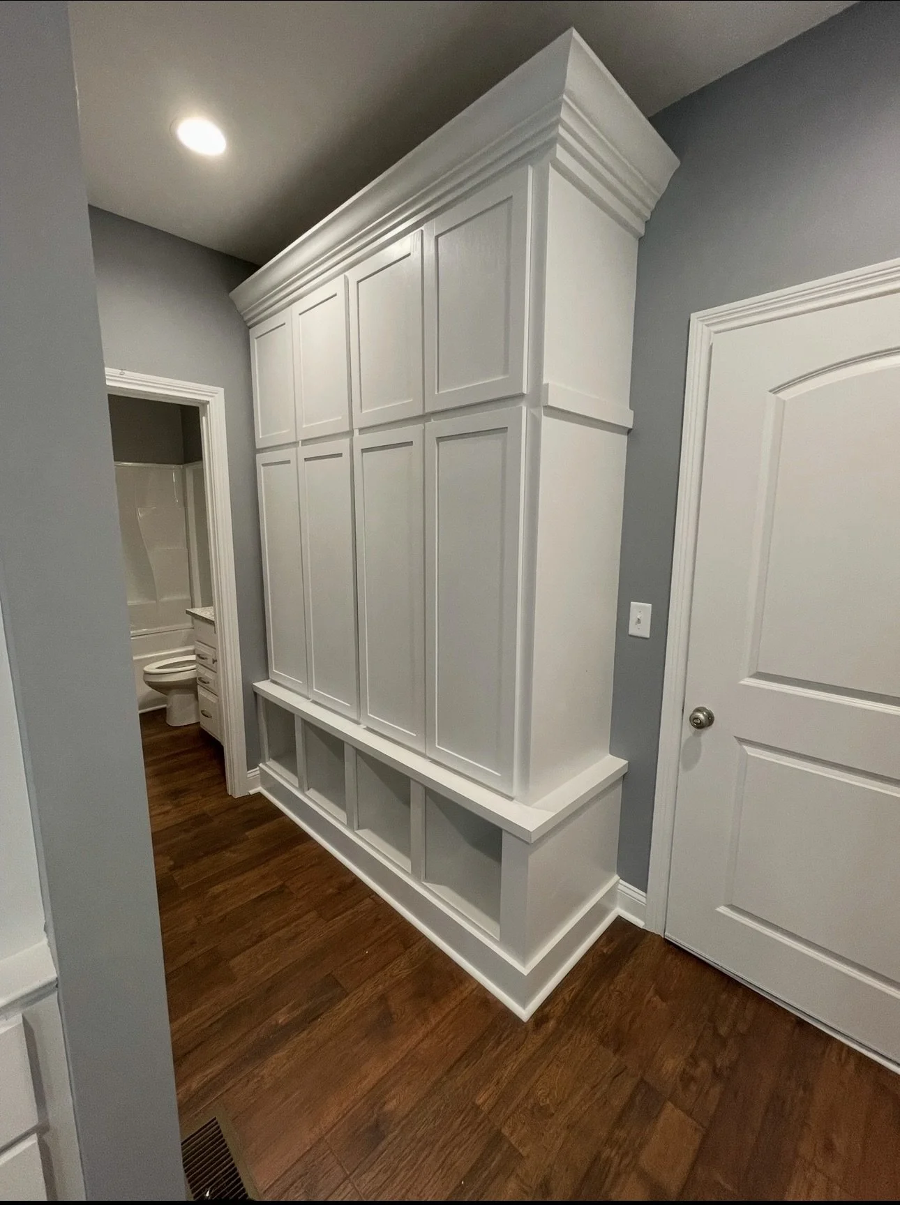White built-in cabinet with closed doors above and open cubbies below, next to a doorway with a room containing a toilet and bathtub visible in the background.