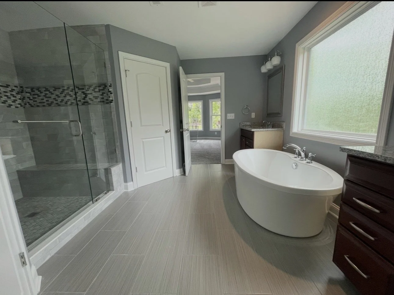 Modern bathroom with a large window, a white freestanding bathtub, a glass-enclosed shower, dark wood cabinetry, and gray tiled flooring.