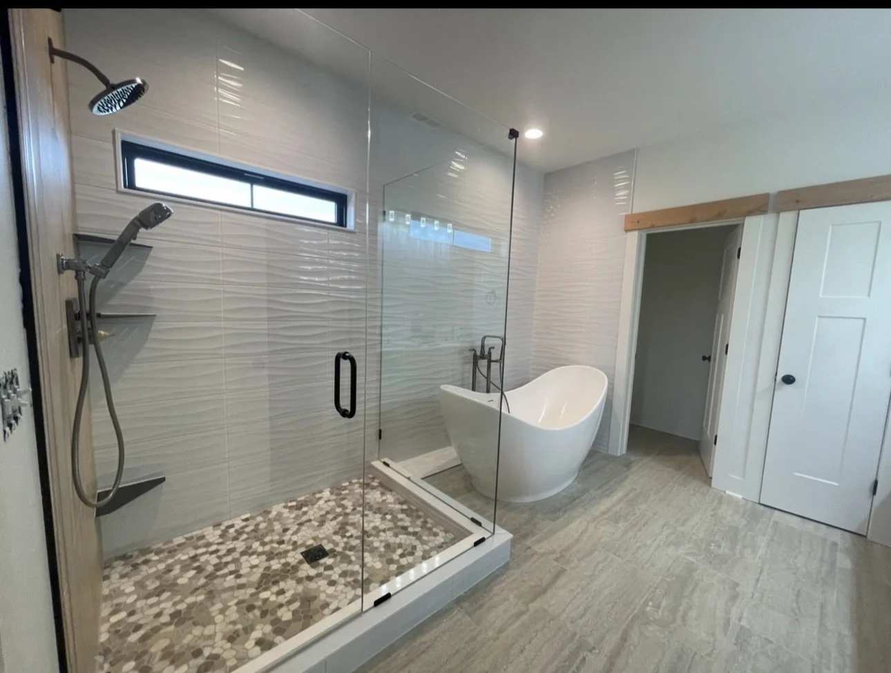 Modern bathroom featuring a glass-enclosed shower area with pebble tile floor, a white freestanding bathtub, a small horizontal window, and wooden trim around the door frame.