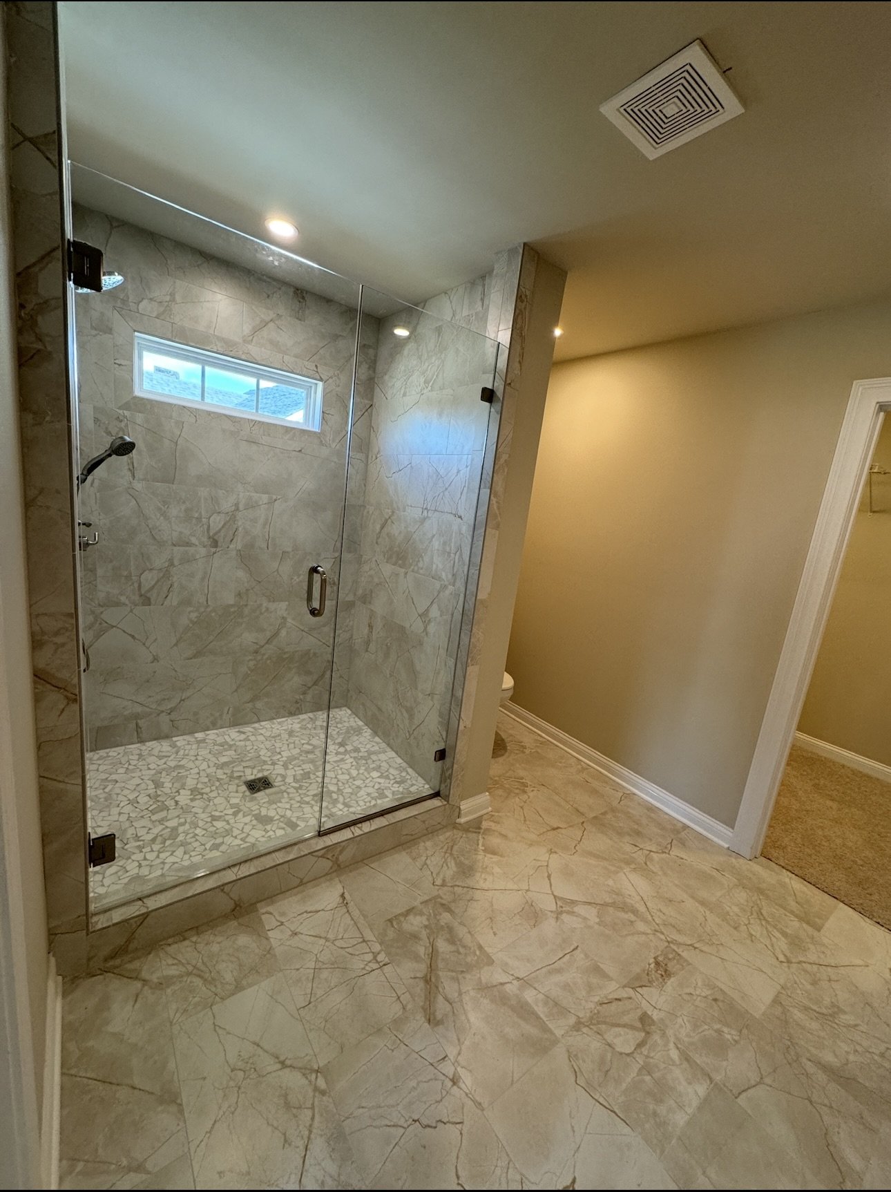 Bathroom with a glass-enclosed shower with beige marble tiles, a small horizontal window near the ceiling, and a beige tiled floor. Part of a toilet is visible behind a beige wall.