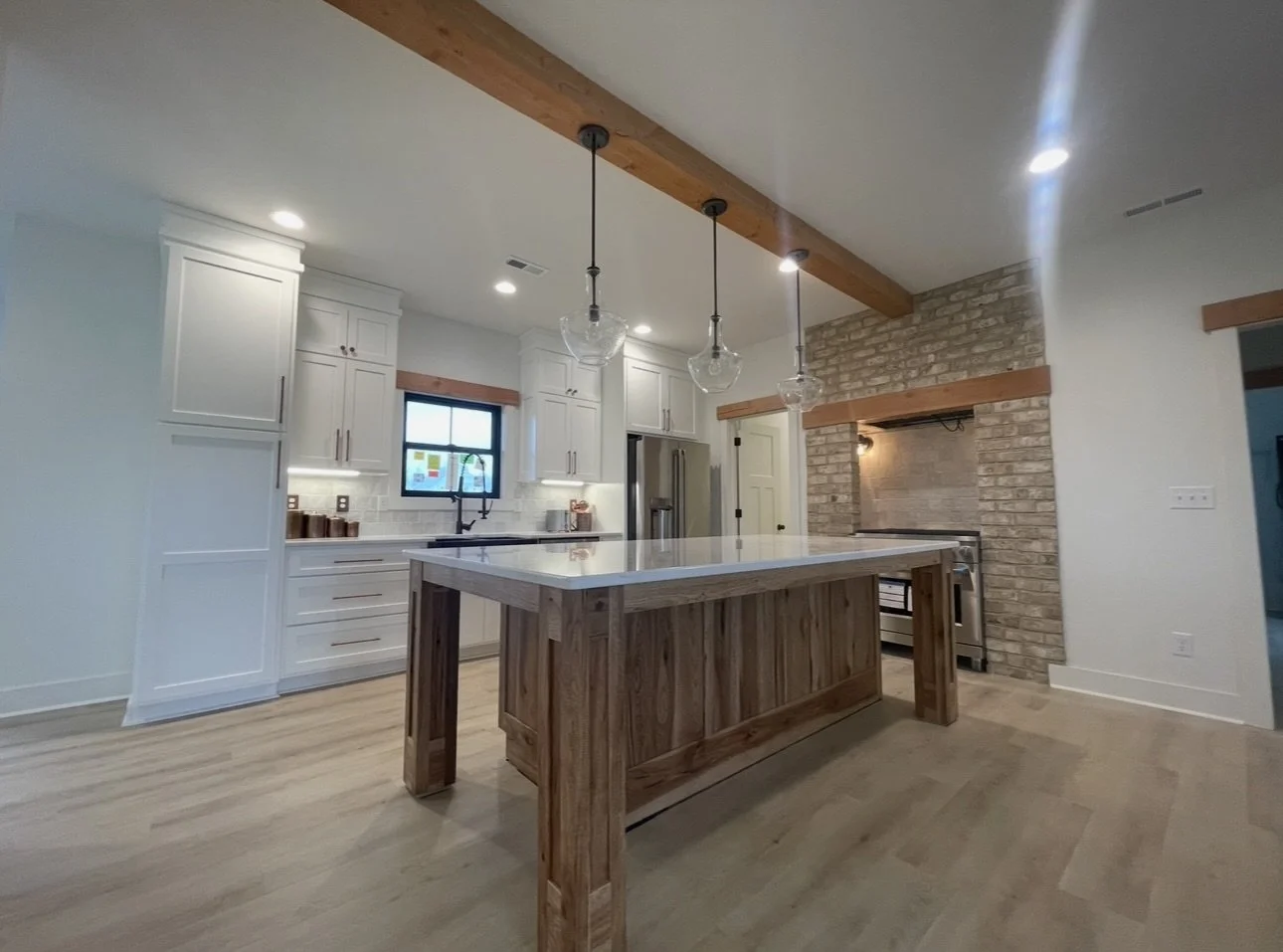 Modern kitchen with white cabinets, a black faucet, a window, stainless steel appliances, brick accent wall, wooden beams, and a wooden kitchen island.