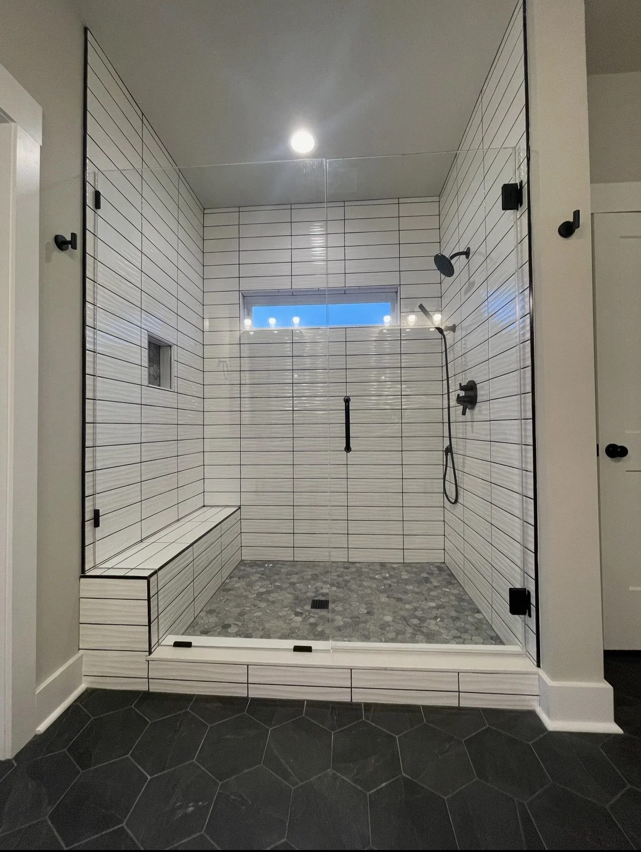 Modern walk-in shower with white tile walls with black grout lines, a small window, built-in bench, pebble stone shower floor, black shower fixtures, and clear glass enclosure.