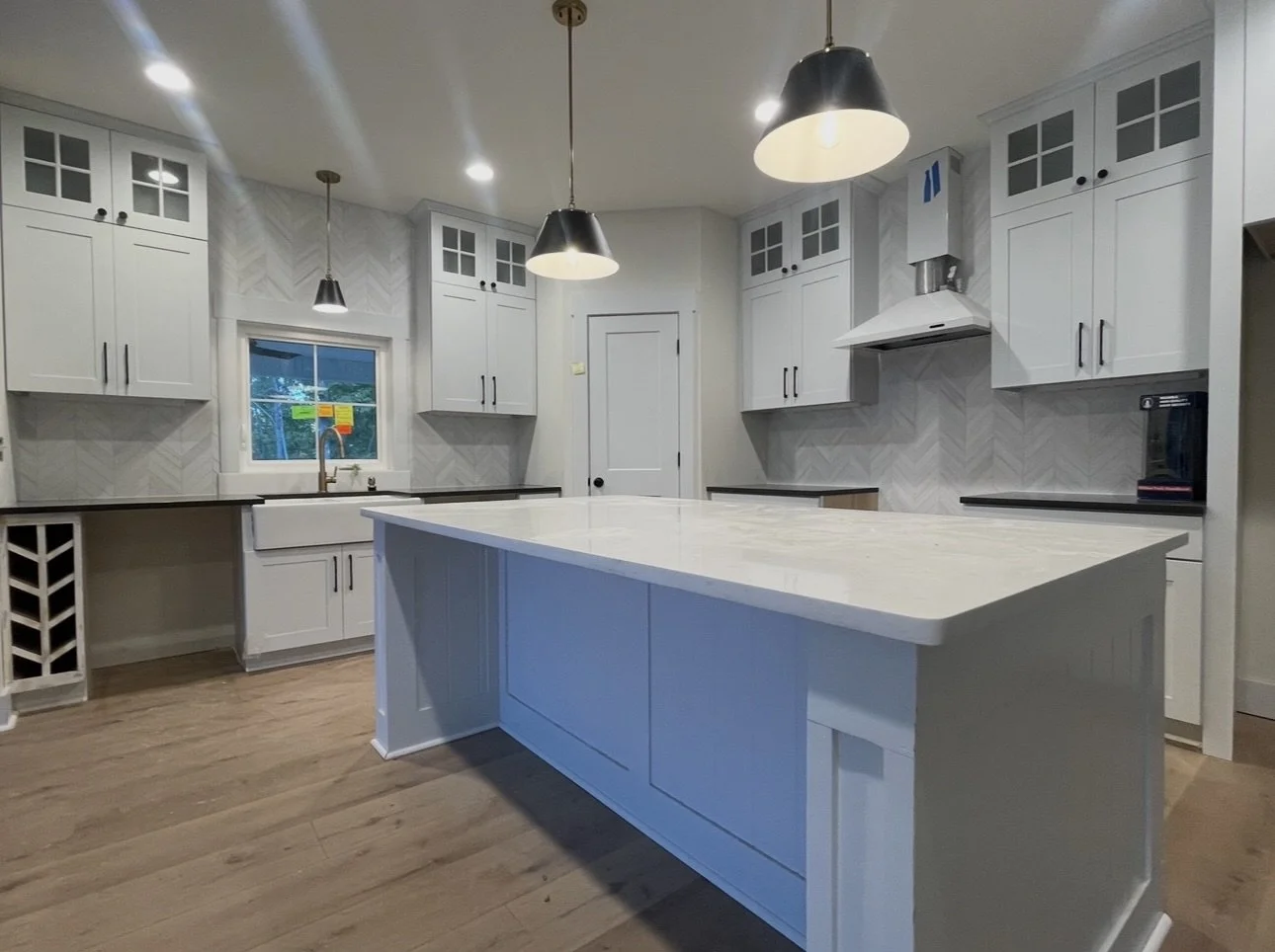 Modern white kitchen with a large island, white cabinets with glass doors, a window above the sink, black hardware, pendant lights, and a wood floor.