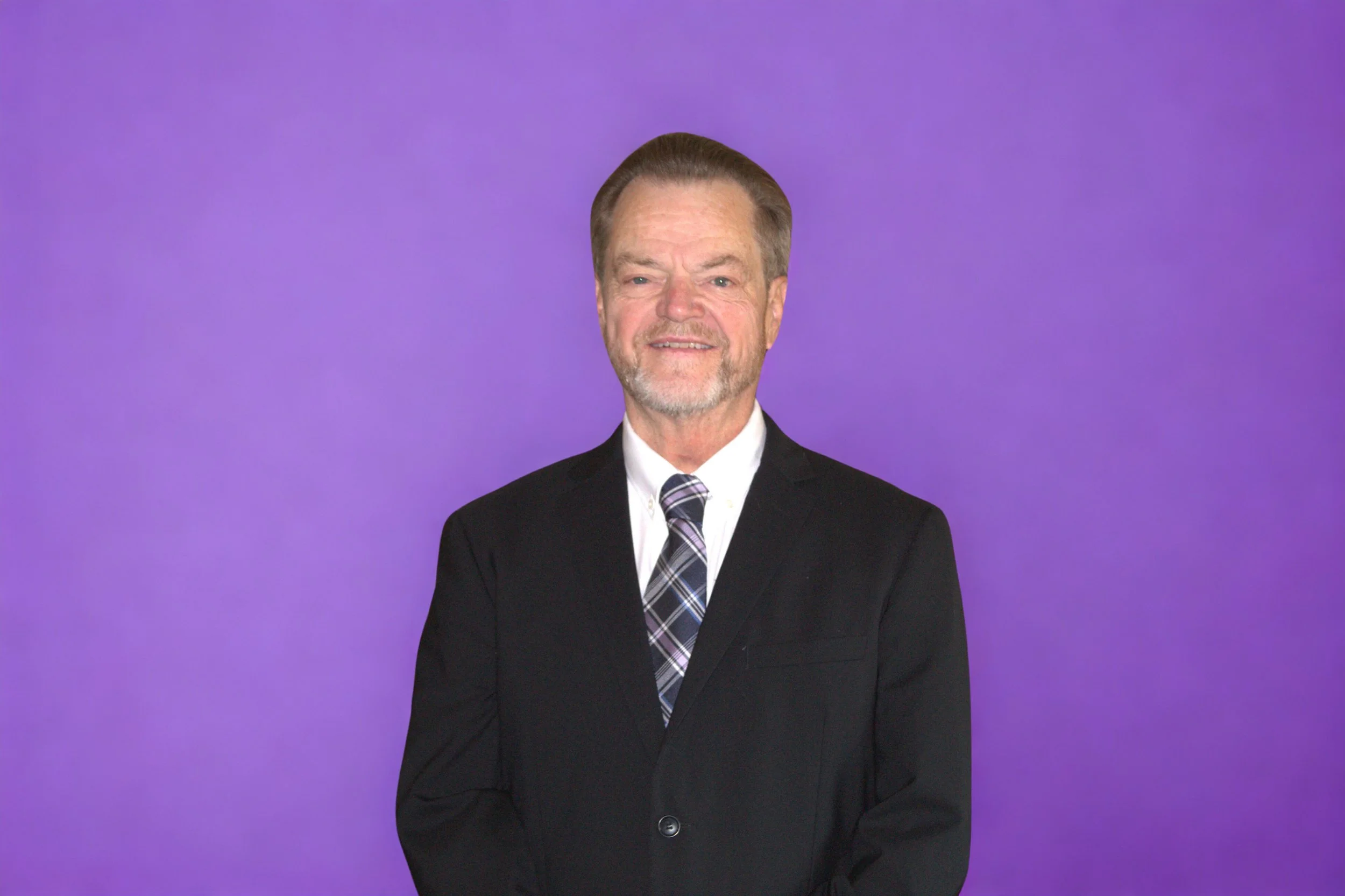 A middle-aged man with gray hair and a beard, dressed in a black suit, white shirt, and patterned tie, standing against a purple background.