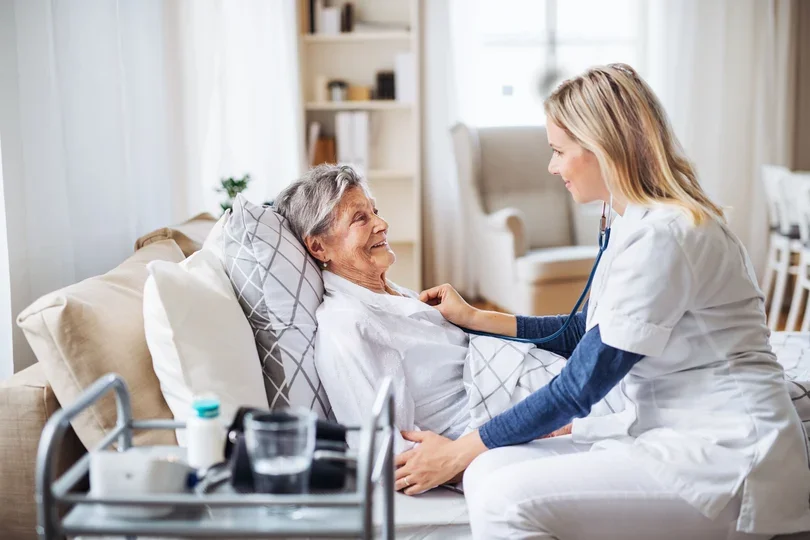 A nurse or caregiver smiling and talking to an elderly woman lying in bed at home, with medical supplies nearby.