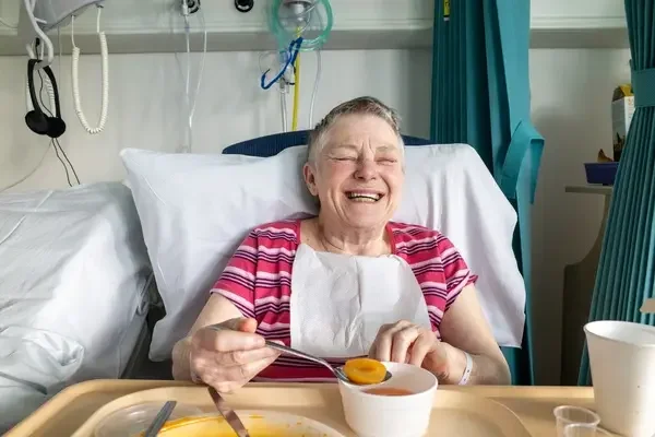 An elderly woman in hospital bed smiling and holding a spoon near her mouth, with food on a tray in front of her.