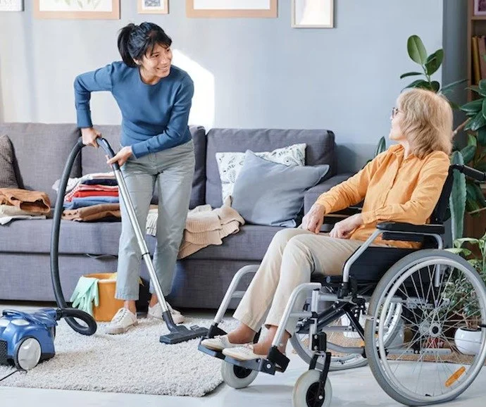 A woman with short dark hair in a blue sweater and gray pants is vacuuming a gray carpet in a living room, talking to an older woman with shoulder-length blonde hair in an orange shirt and beige pants sitting in a wheelchair.