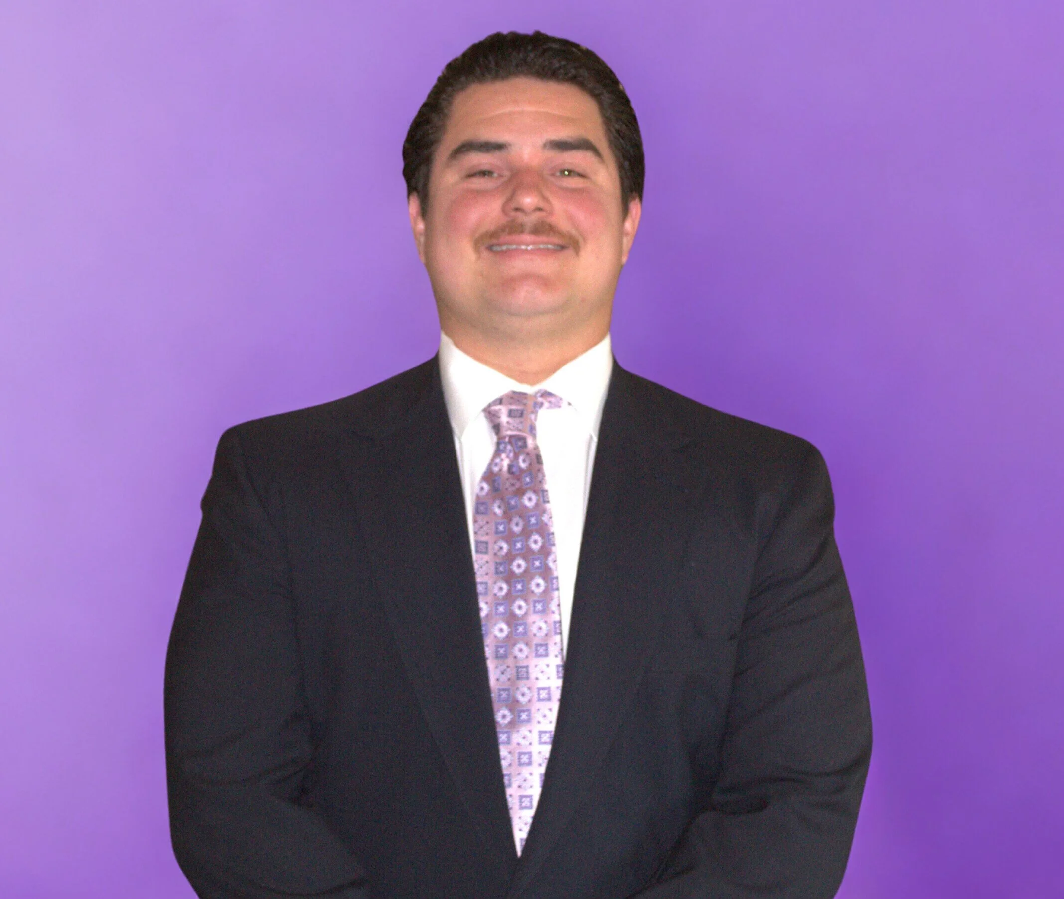 A man with brown hair and a mustache smiling, wearing a suit and tie with a purple patterned design, standing against a purple background.