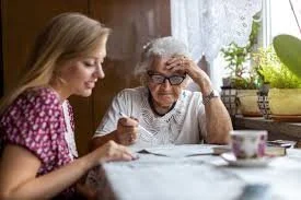 A young woman and an older woman sit at a table in a cozy kitchen, smiling and talking, with a teacup and a plant on the windowsill.