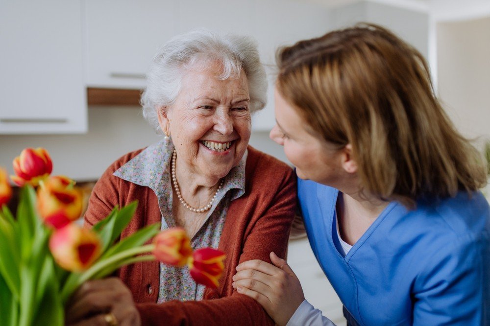 A senior woman smiling and holding a bouquet of flowers, engaging warmly with a caregiver in a blue uniform in a bright kitchen setting.