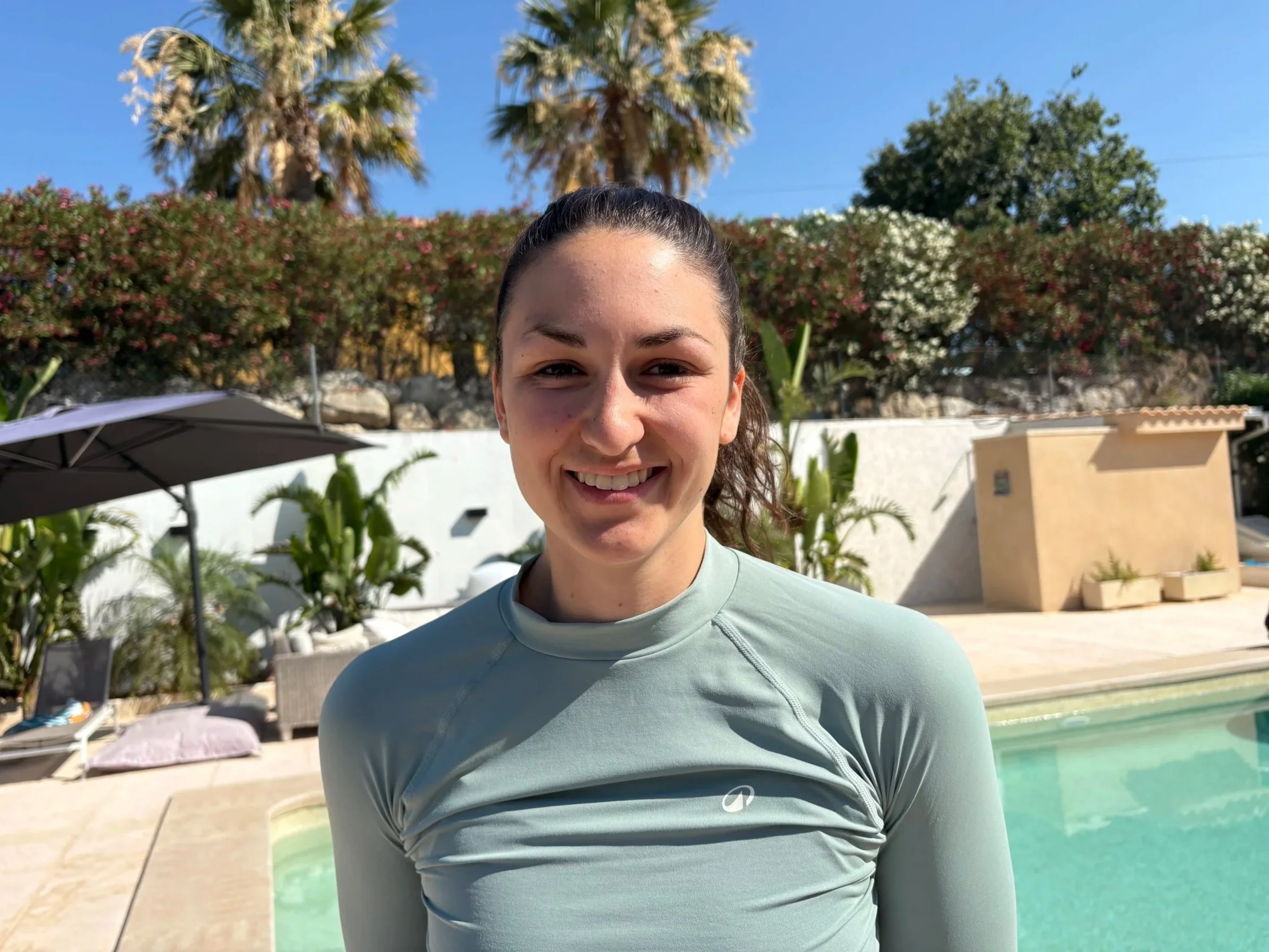 Jeune femme souriante en tenue de sport près de la piscine à l'extérieur, arrière-plan avec plantes et arbres, ciel bleu.
