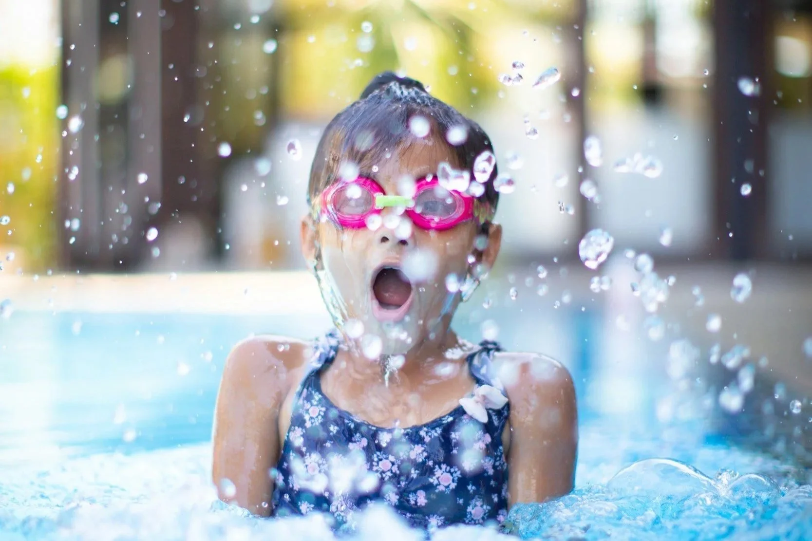 Une jeune fille nageant dans une piscine, portant des lunettes de natation roses, avec une bouche ouverte d'émerveillement ou de surprise, entourée de bulles d'eau.