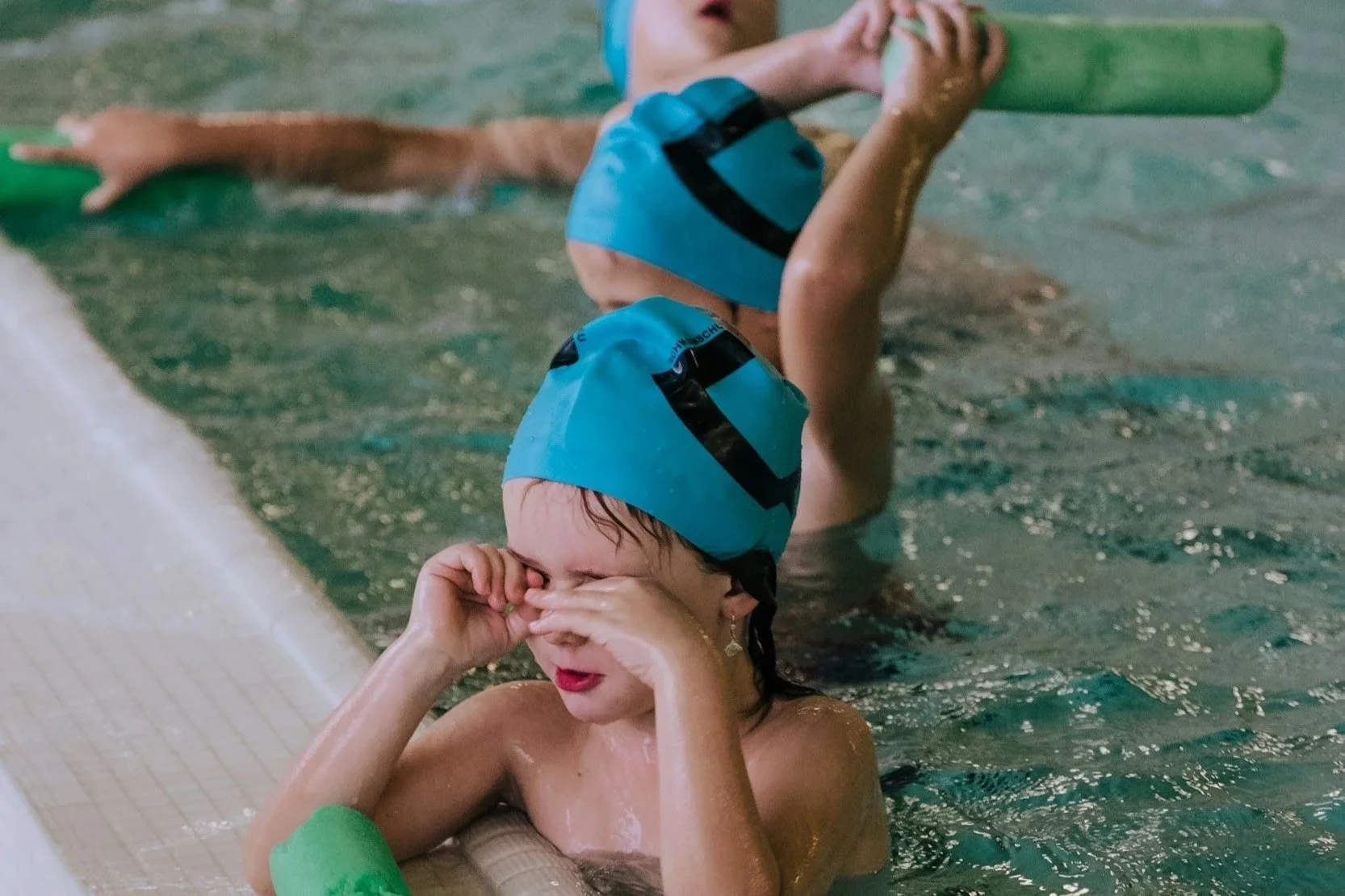Deux enfants en maillot de bain et bonnet de bain bleus, s'apprêtant à faire de la natation, dans une piscine intérieure.