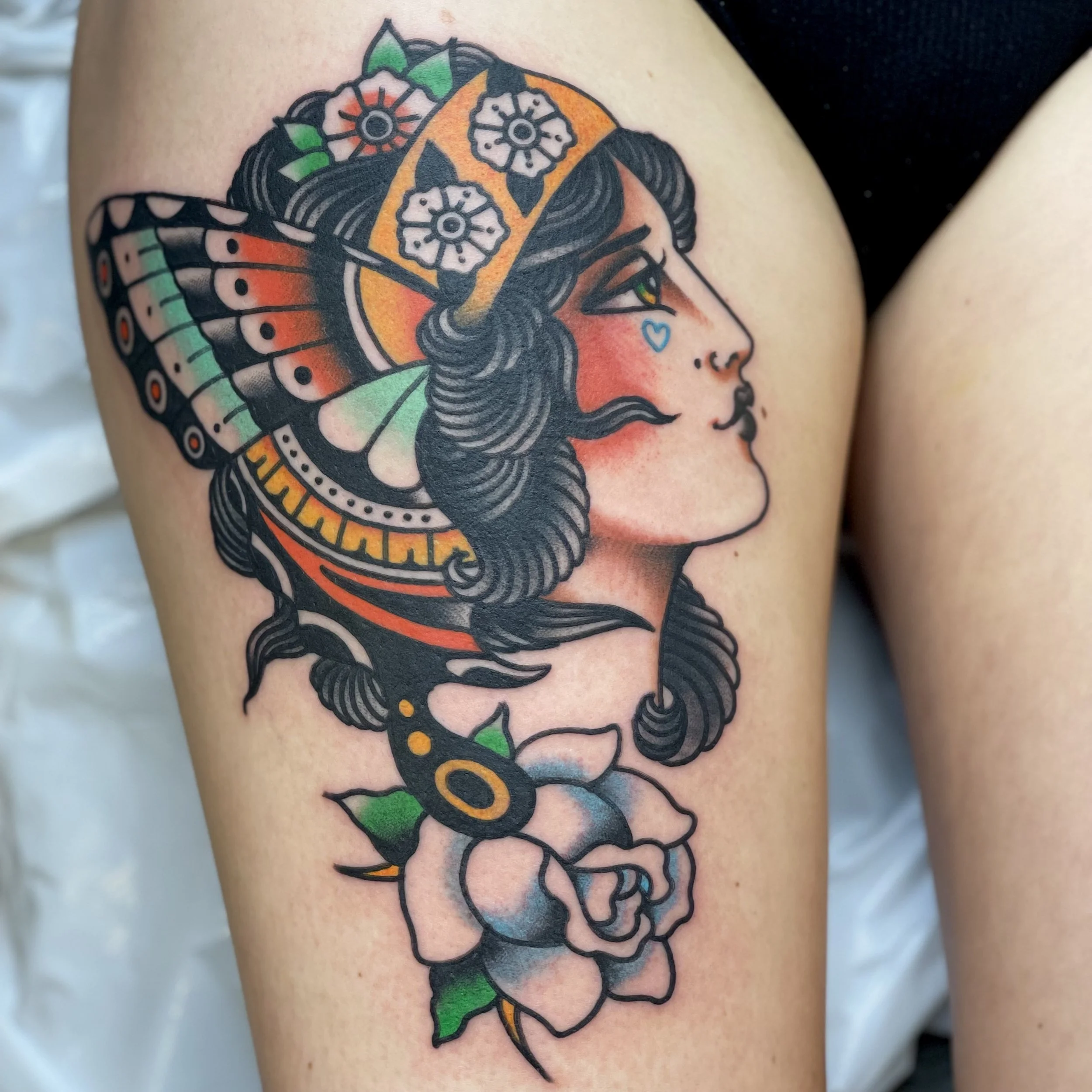 Tattoo of a woman with dark curly hair, wearing a yellow and white floral headband, with a butterfly wing accessory, a small blue heart tattoo near her eye, and a large white rose at the base.