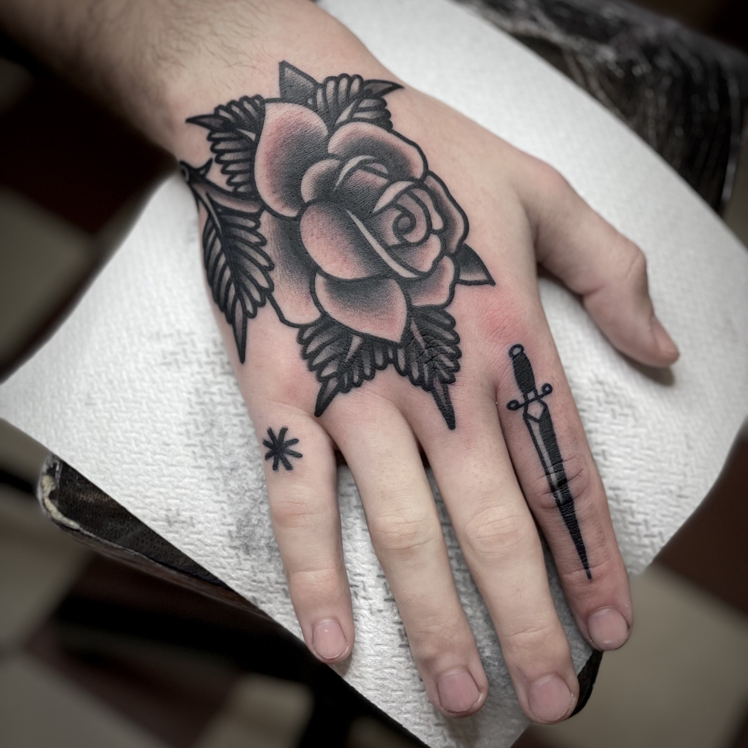 Tattoo of a rose with leaves on the hand, and a dagger tattooed on the index finger.