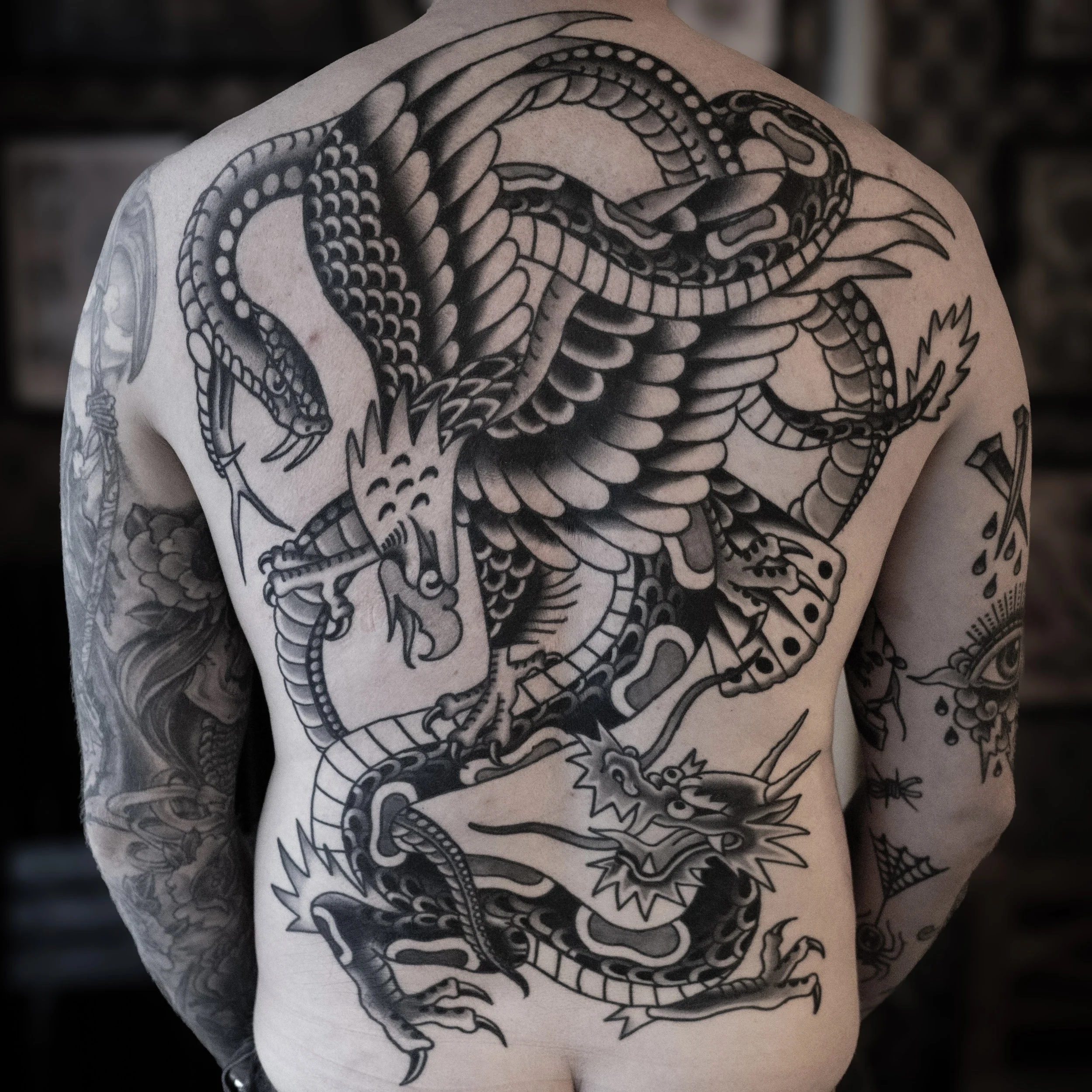 Back tattoo of a large, detailed cartoon-style dragon with an open mouth, sharp claws, and intricate scales covering the person's back, with additional tattoos on the arms.