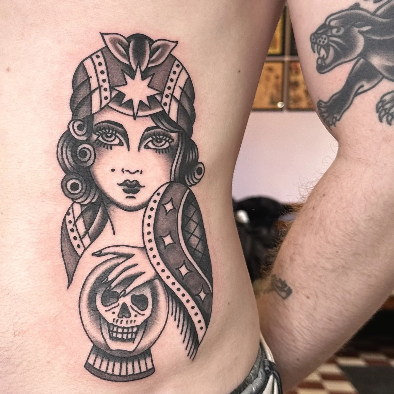 Tattoo of a woman's face with large eyes and dark hair, wearing a headscarf, holding a skull with a dagger through it.