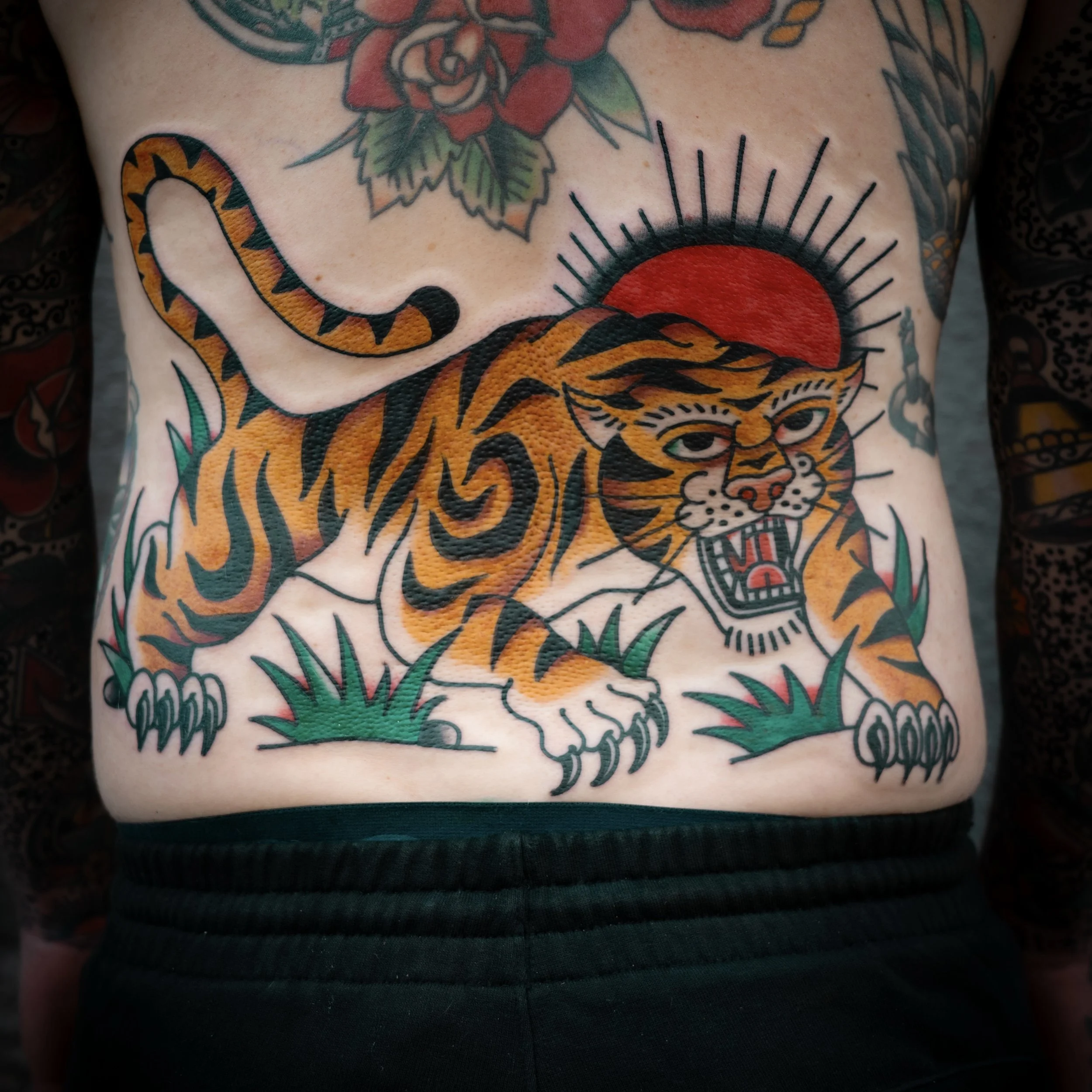 Tattoo of a fierce tiger in a traditional style, with black and orange stripes, sharp teeth showing, and green grass under its paws. The tiger's head is positioned behind a red sun with black rays radiating outward, and the tattoo is located on a per