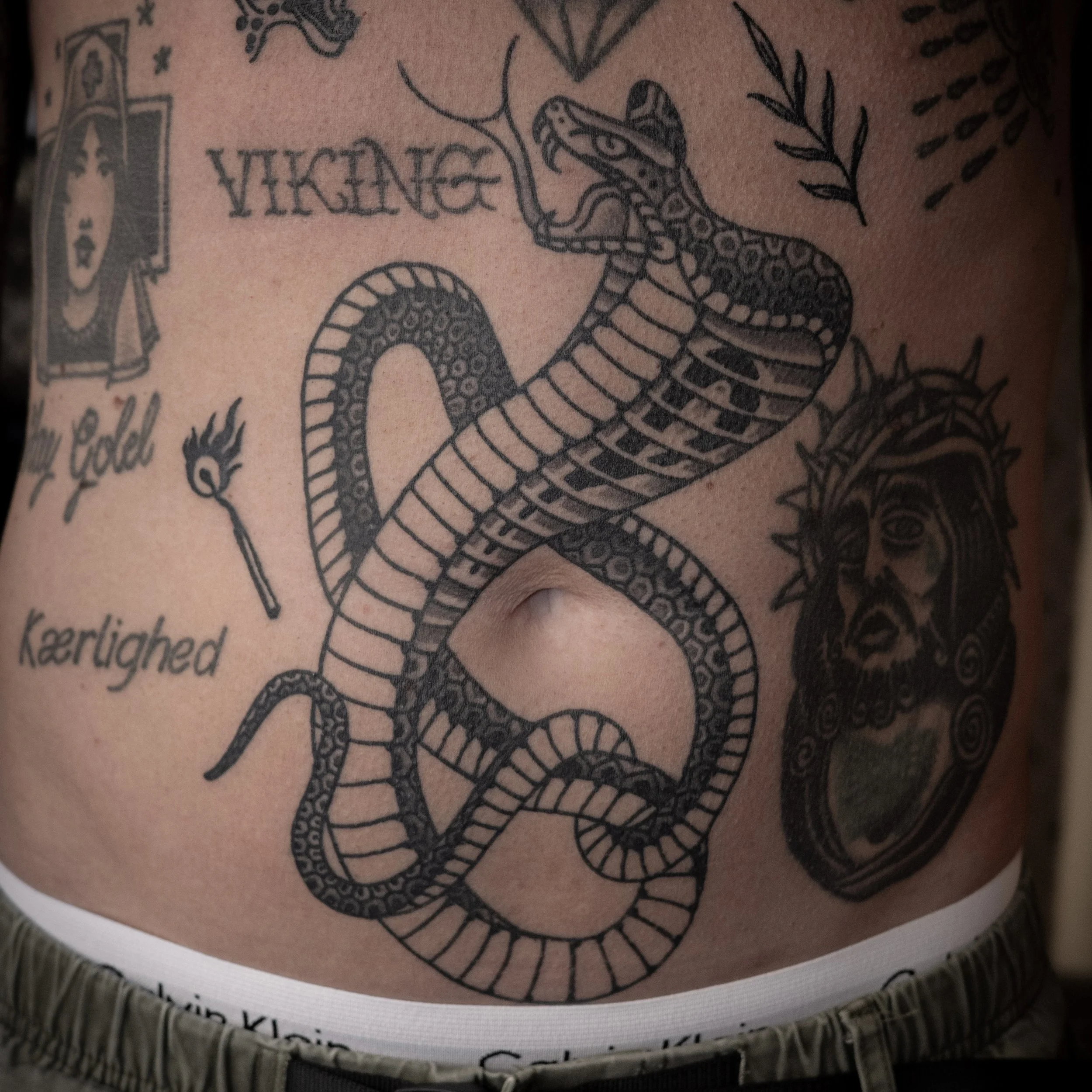Tattoo on person's abdomen featuring a snake with a forked tongue, a crowned head with an open mouth, and various words like 'VIKING,' 'gold,' and 'Kærlighed'.