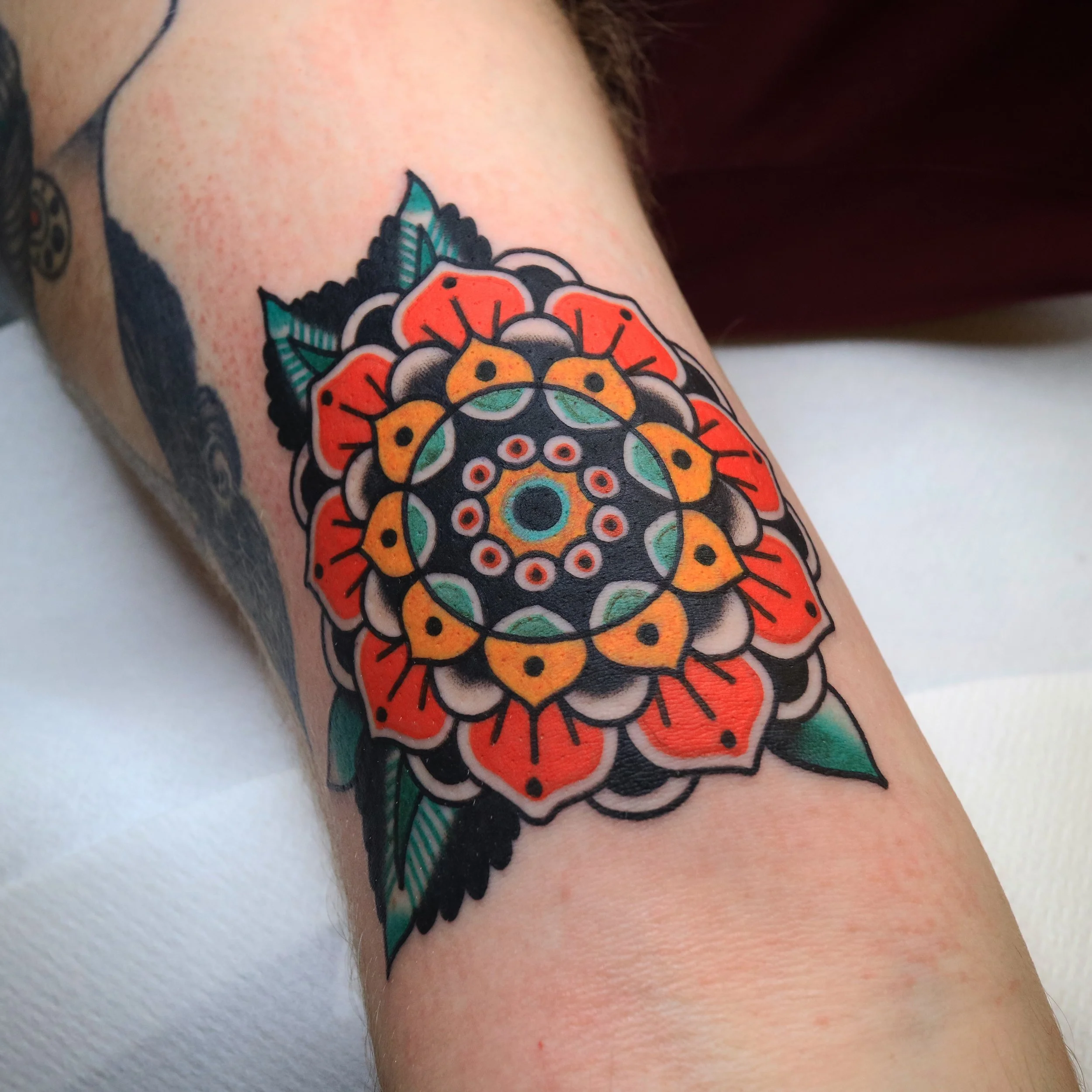Colorful mandala-style tattoo on person's arm featuring a central circle with multiple rings of geometric and floral patterns in black, orange, yellow, green, and red, with surrounding leaves.