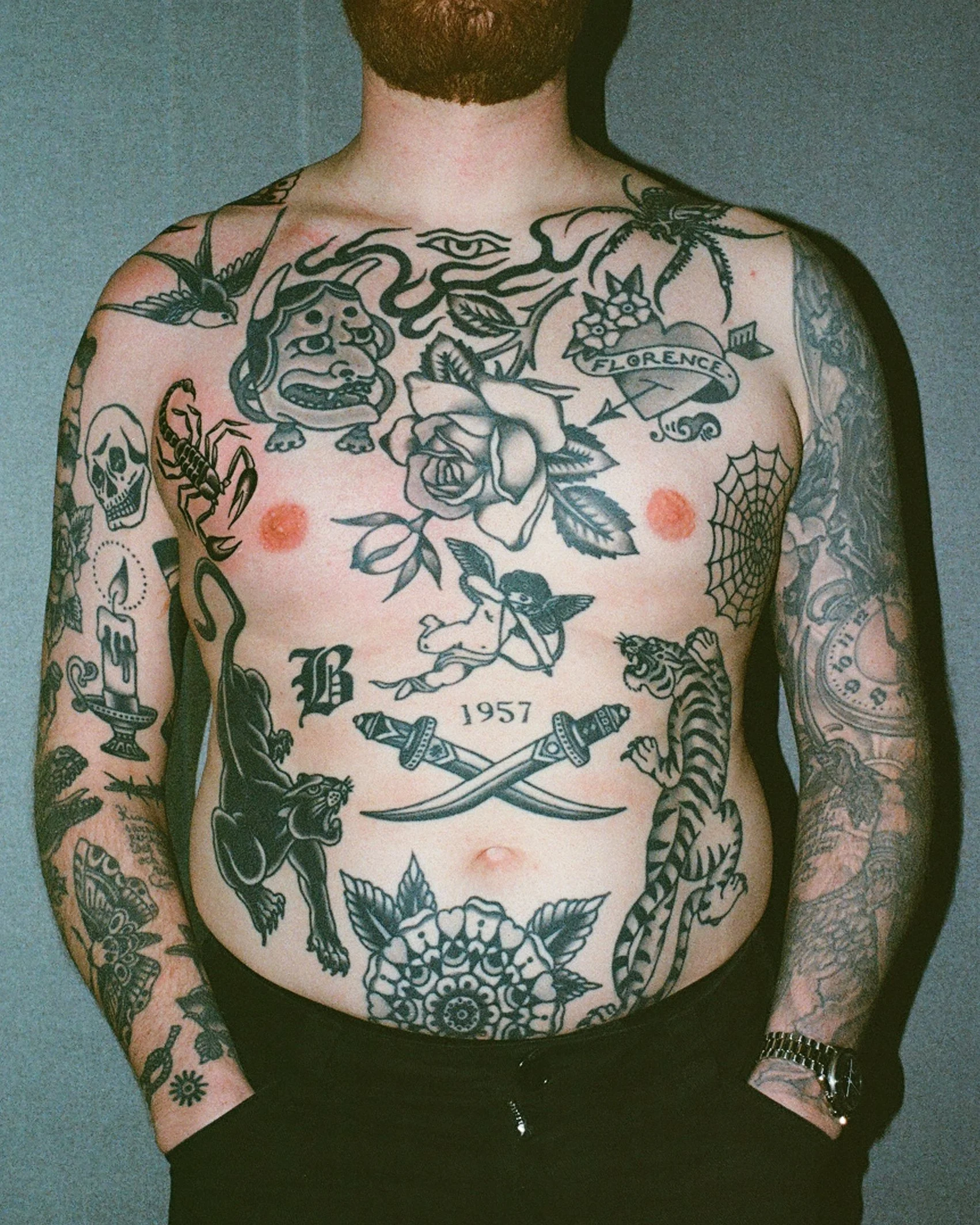 A shirtless man with numerous black tattoos on his chest and arms, including a rose, a cherub, a tiger, a spider web, and skulls, standing against a plain background.