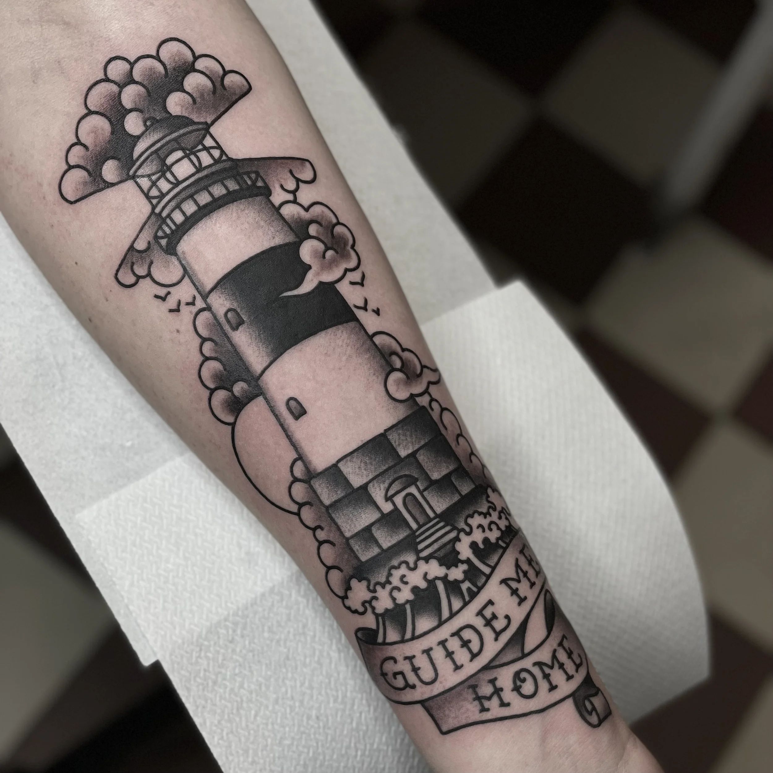 Tattoo of a lighthouse surrounded by clouds and birds with a banner that reads 'Guide Me Home' on a person's arm.