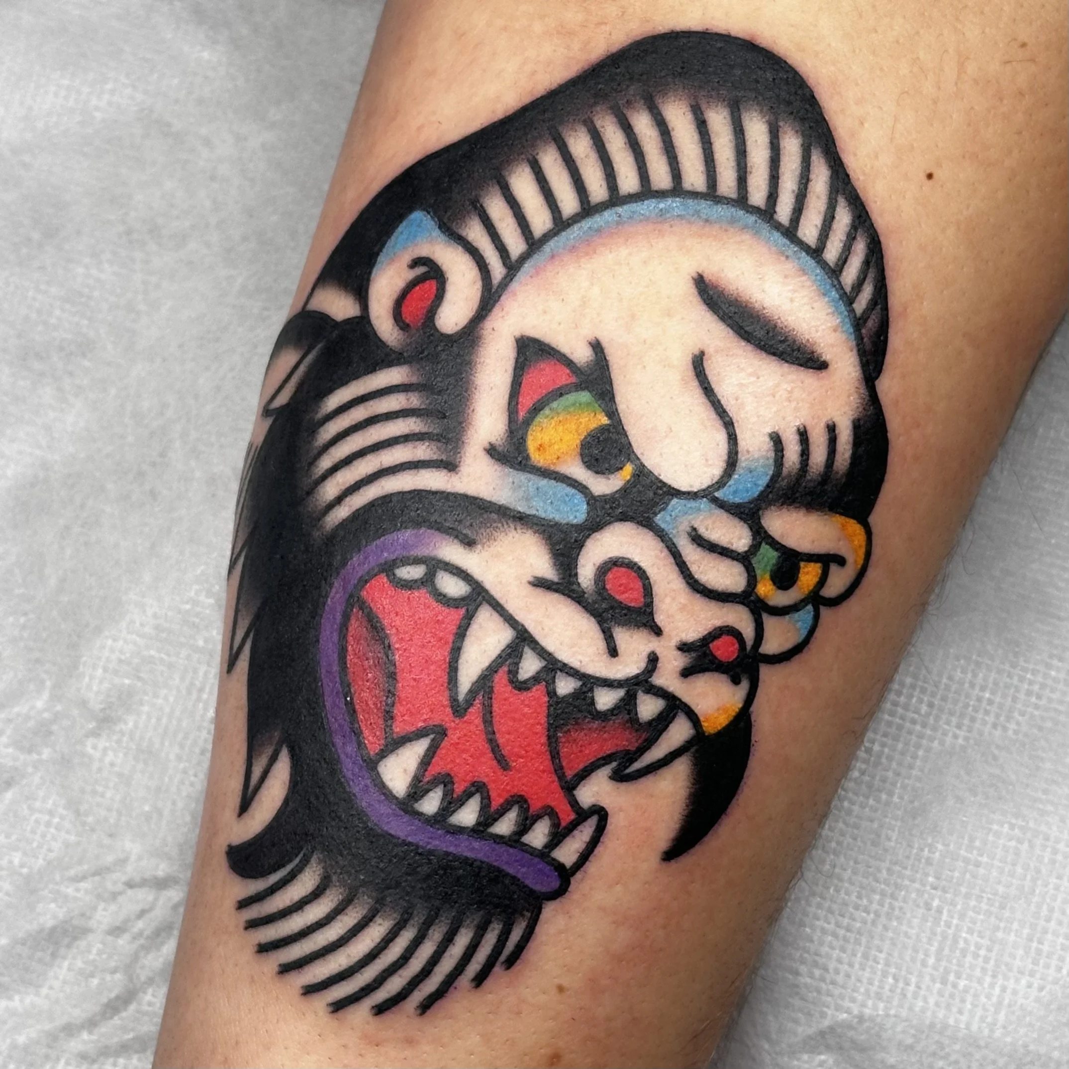 Traditional-style tattoo of a stylized dragon skull with bold black outlines and colorful accents, including red, yellow, green, and purple.