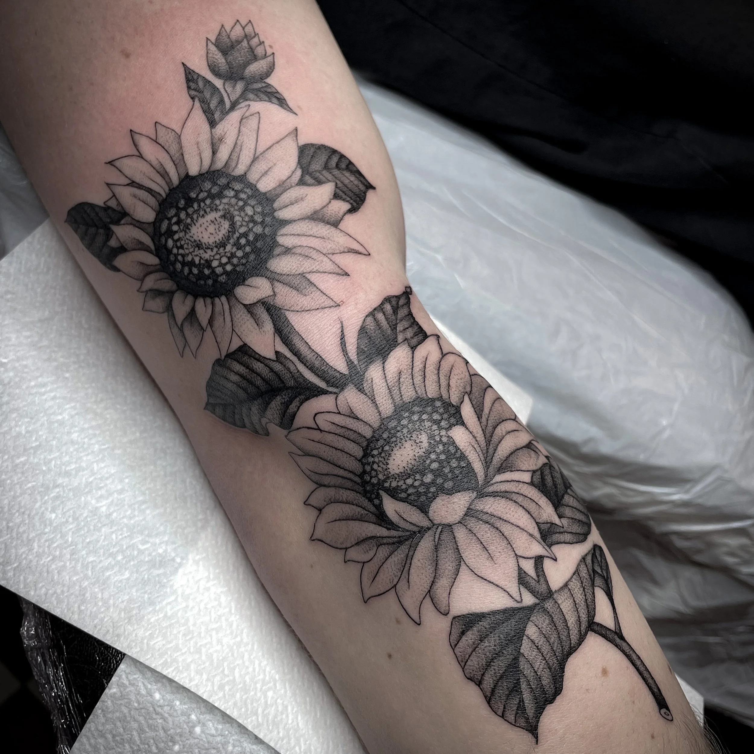 Black and gray tattoo of two sunflowers with detailed petals, leaves, and stems on a person's forearm.