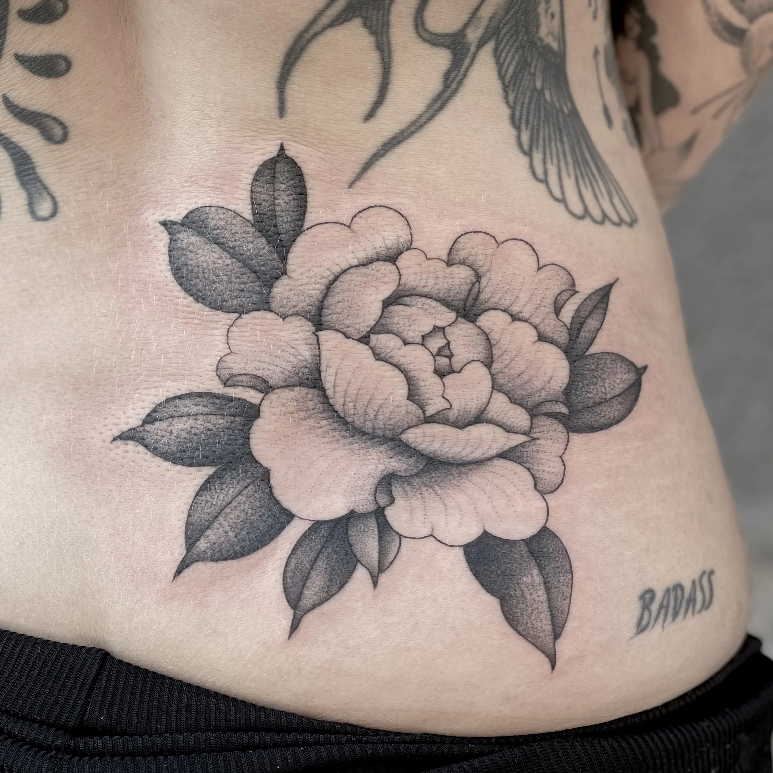 Black and gray tattoo of a blooming peony flower with leaves on person's side.