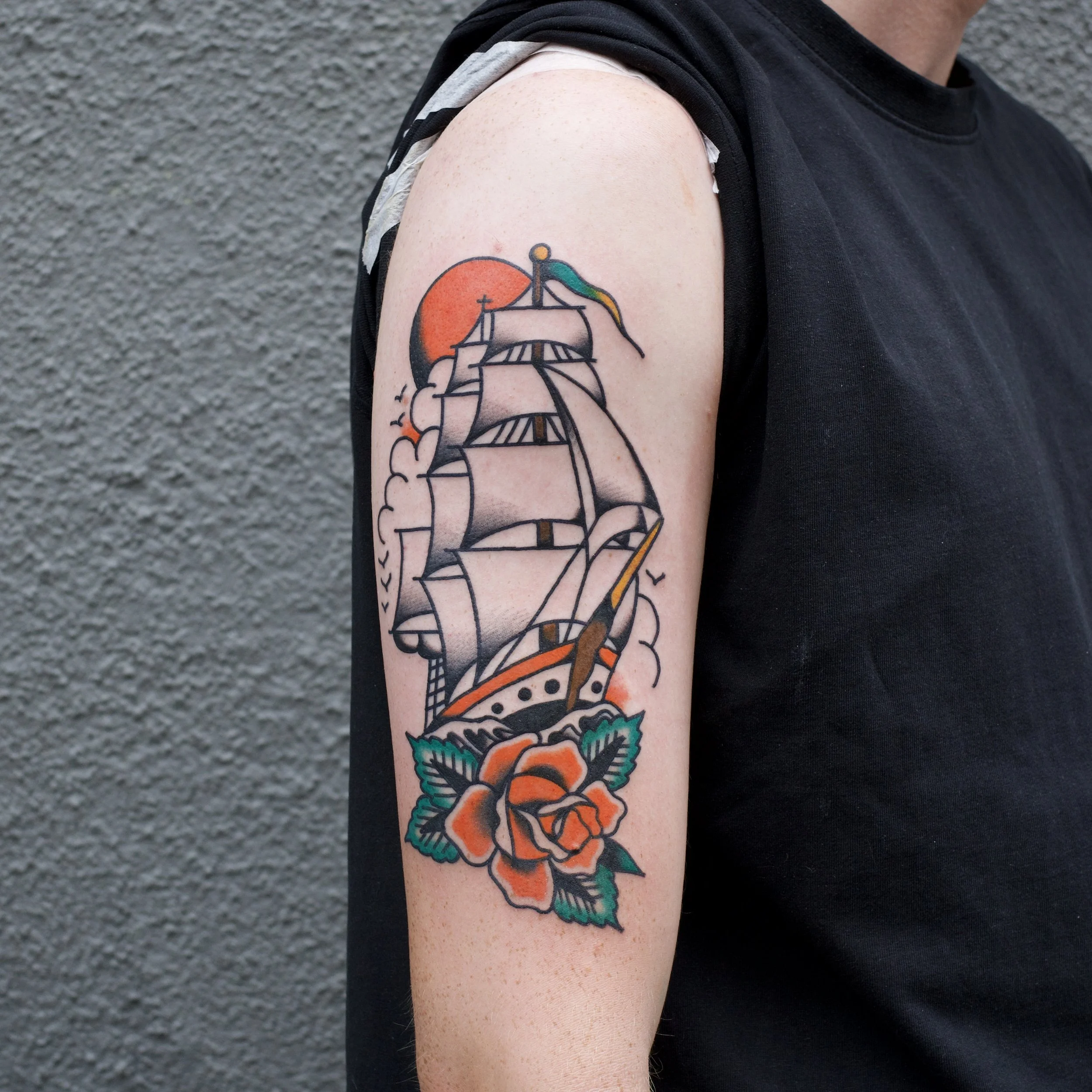 Tattoo of a sailing ship with orange sun, flying flag, and a rose at the bottom on person's upper arm.