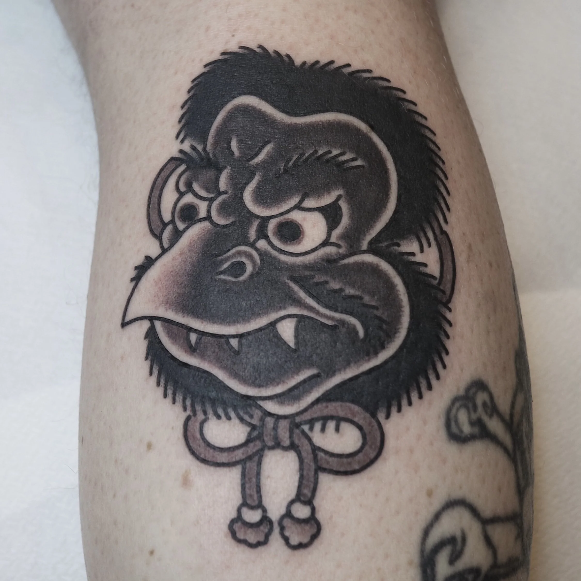 Tattoo of a cartoonish, angry-looking gorilla face with exaggerated features, a prominent nose, and a bow tied around its neck, on a person's arm.