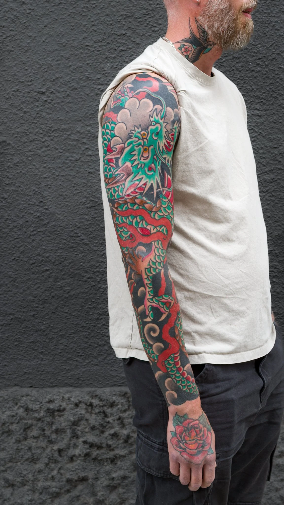 A man with gray hair and a beard displaying a colorful Japanese-style dragon tattoo sleeve on his right arm, standing against a dark textured wall.
