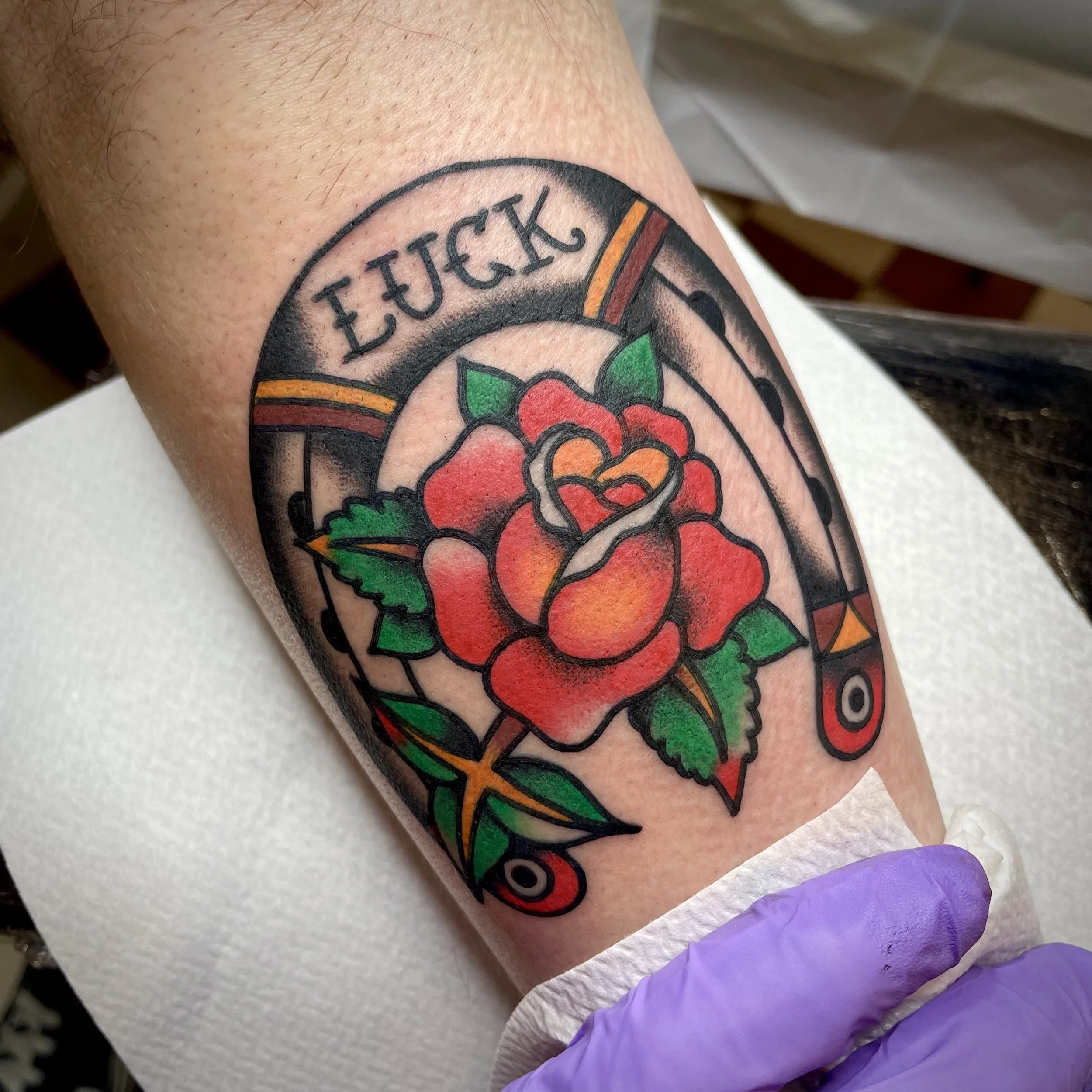 A colorful traditional-style tattoo of a red rose with green leaves, encircled by a banner that reads 'LUCK'. The tattoo also features black lines and bold colors, including yellow and orange accents.