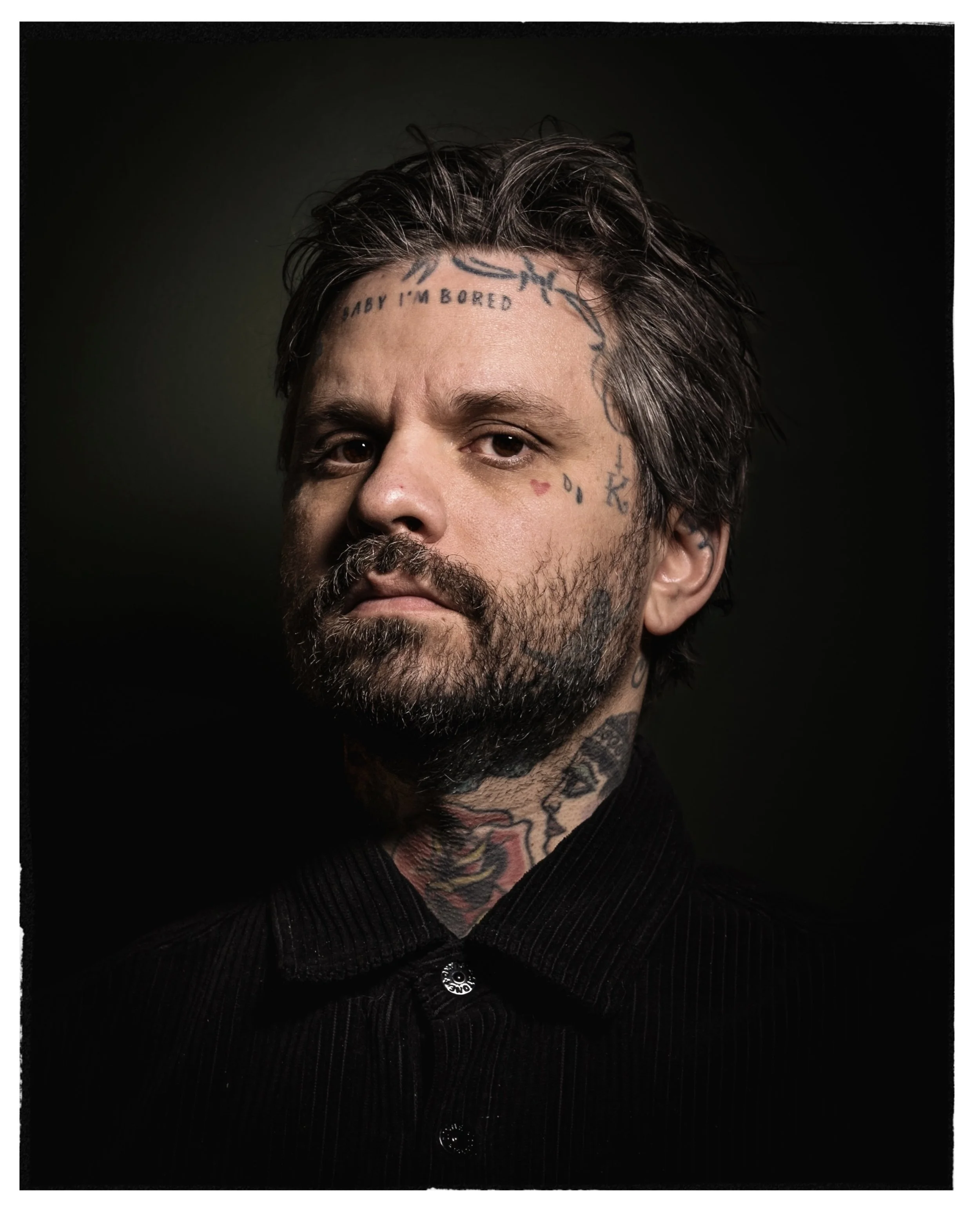 Close-up portrait of a man with dark hair, beard, and numerous tattoos on his face, neck, and head, wearing a black collared shirt, against a dark background.