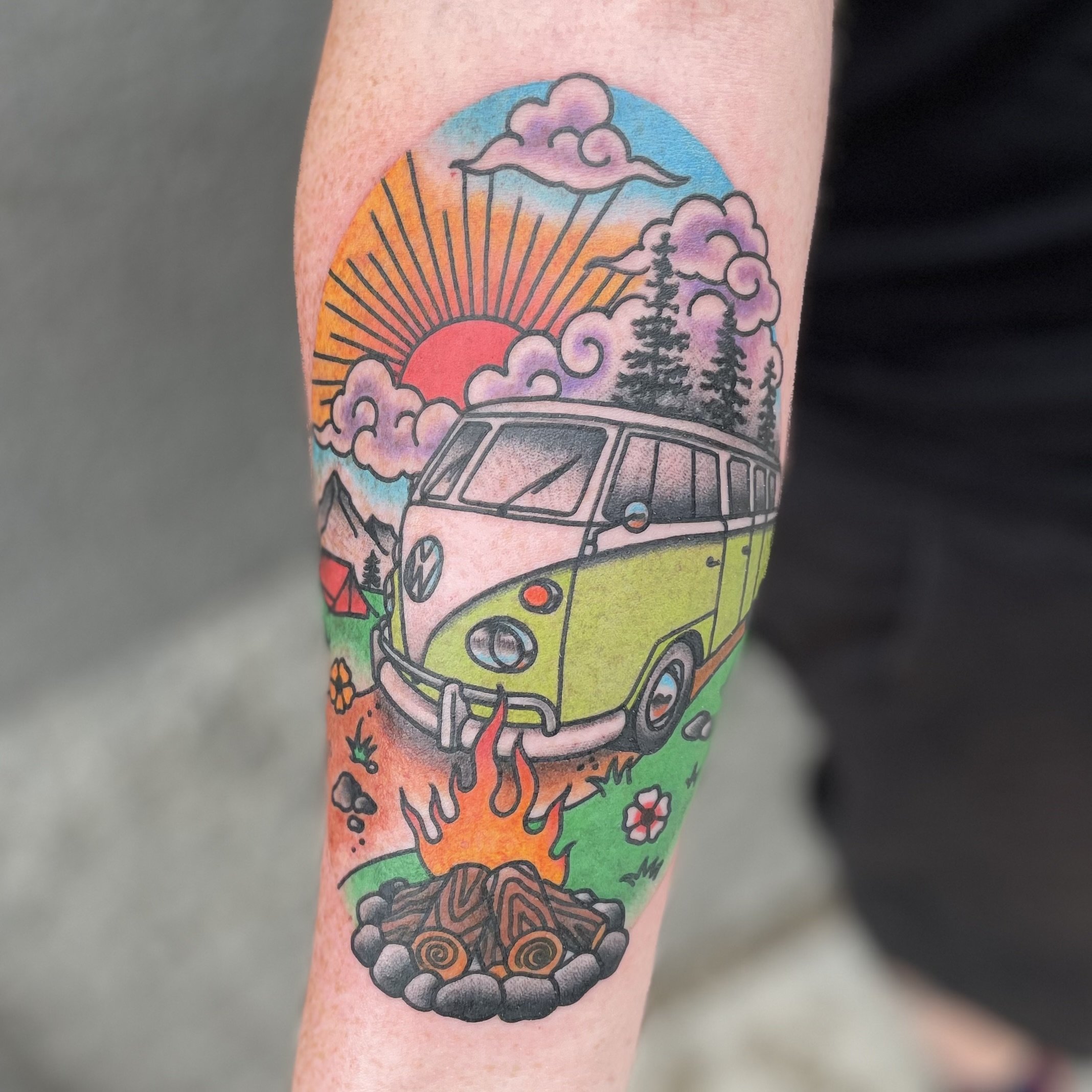 Colorful tattoo of a vintage Volkswagen van parked by a campfire with mountains, pine trees, a tent, and a sunrise in the background.