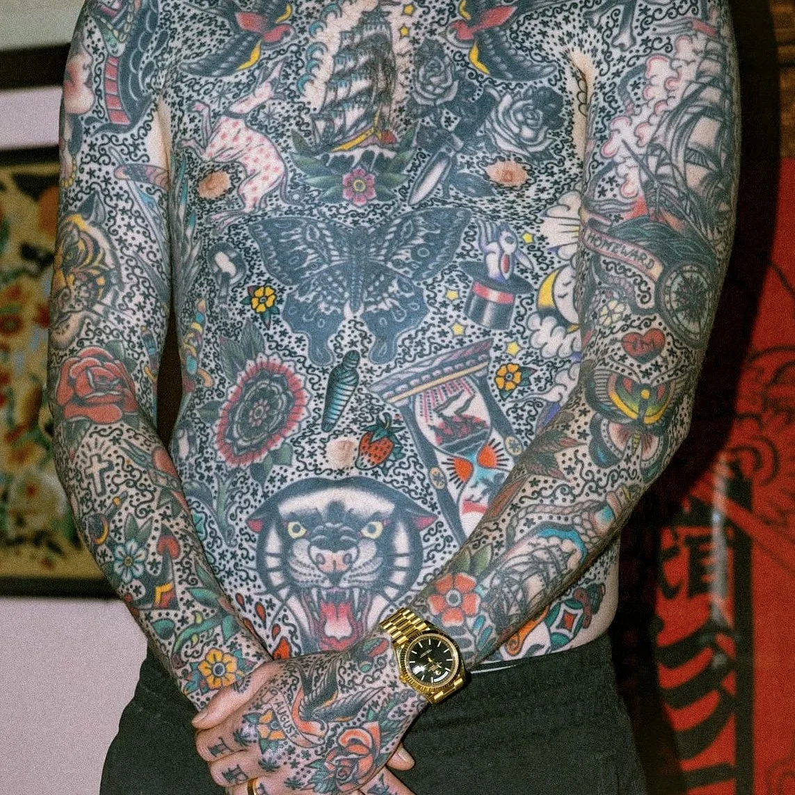 A person with extensive colorful tattoos on their torso and arms. The tattoos include butterflies, roses, a tiger's face, an hourglass, a top hat, strawberries, and various other designs. The person is wearing a gold wristwatch and black pants.