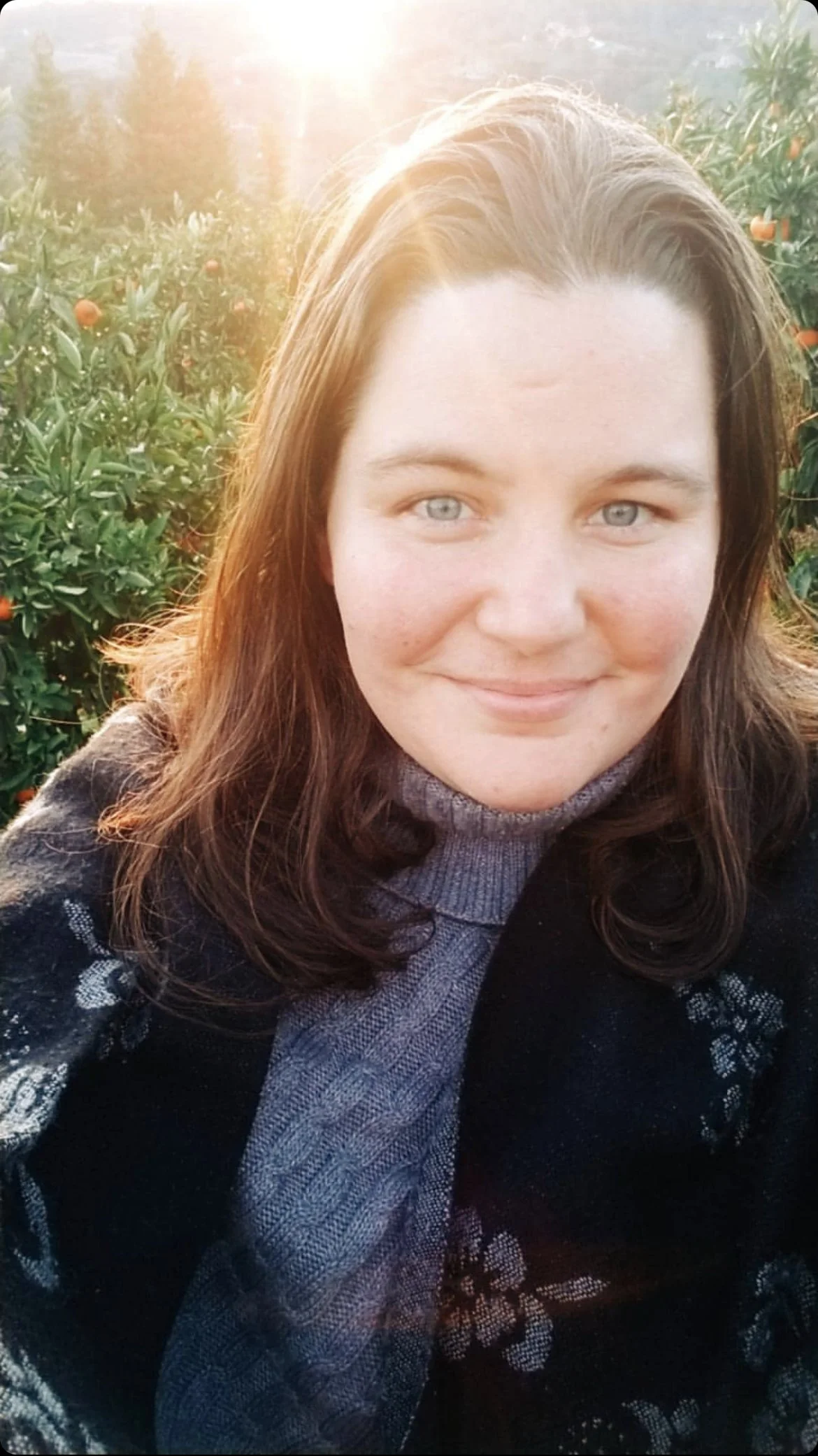 A young woman with light skin and blue eyes smiling outdoors in a field of orange trees during the daytime, with sunlight shining in the background.