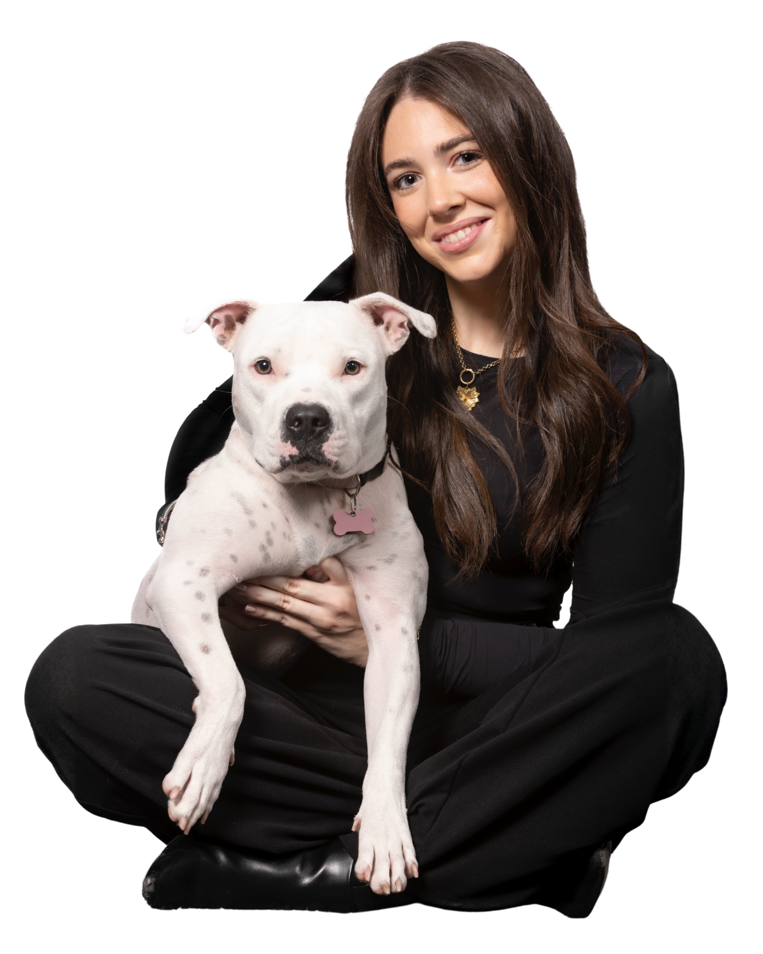 A woman with long wavy brown hair wearing a black long-sleeve top and gold necklace, smiling while sitting on the floor and embracing a white dog with a pink bone-shaped collar tag.
