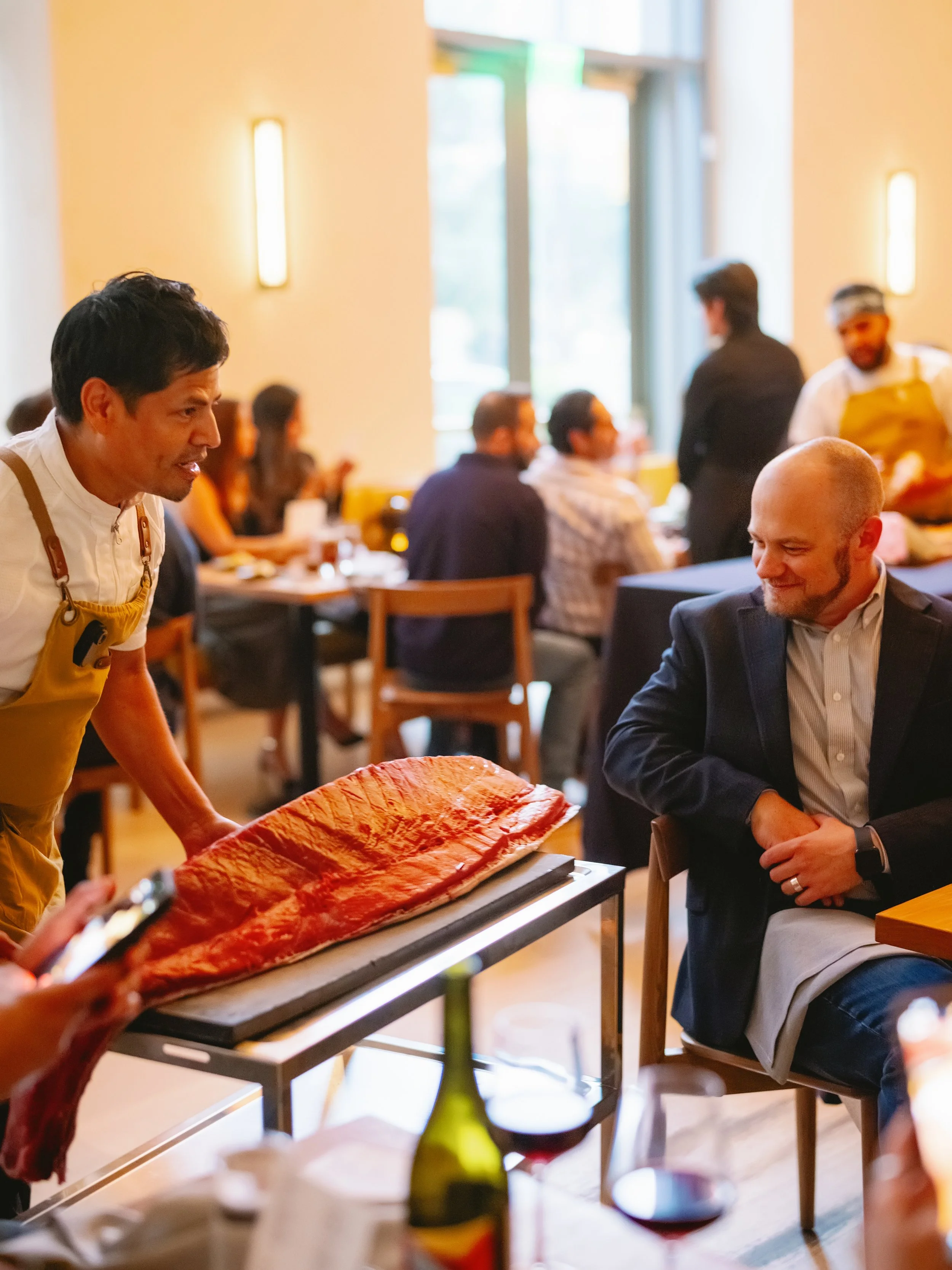 A restaurant scene with a chef serving a large piece of salmon to a seated man in a suit, while other diners enjoy their meals in the background.