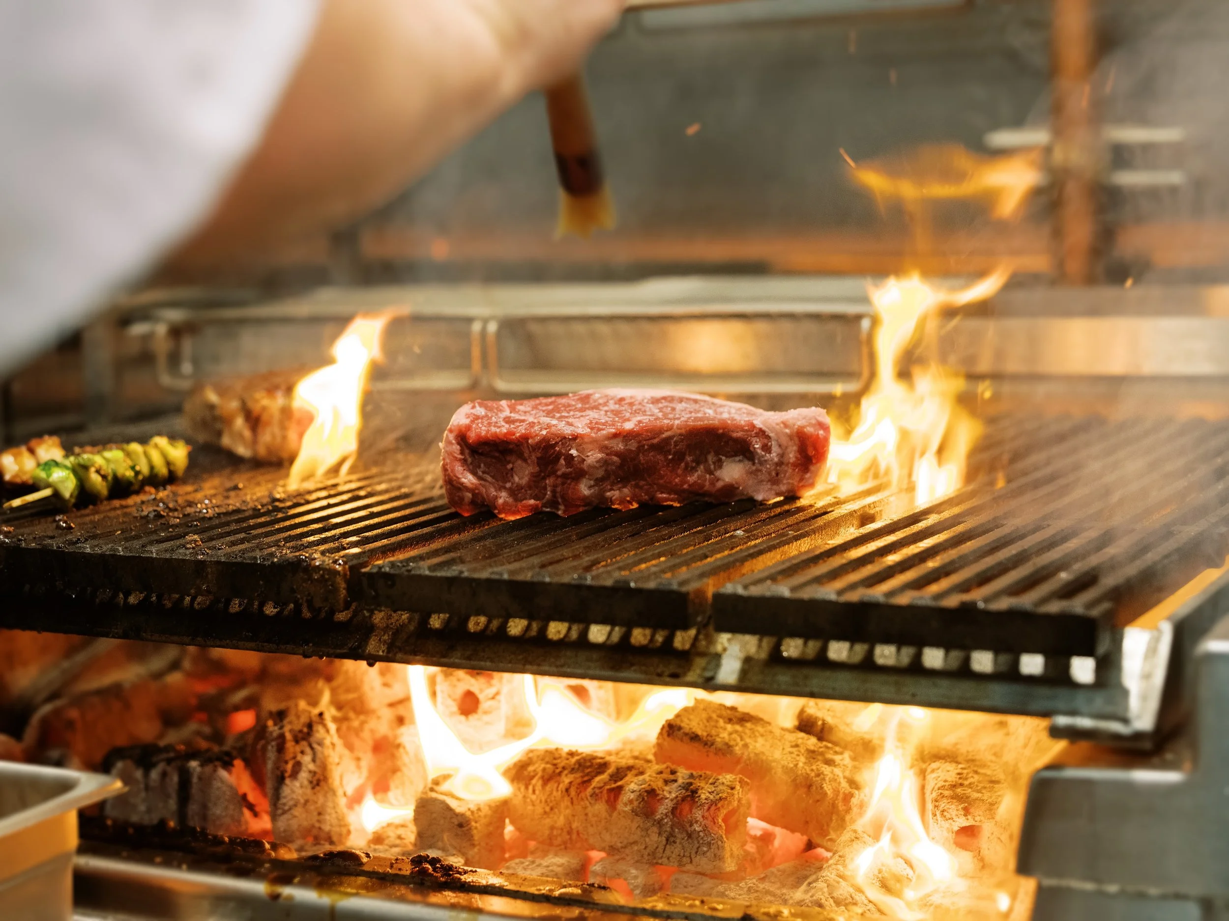 A piece of raw steak cooking on a grill with flames underneath and some green garnishing nearby.