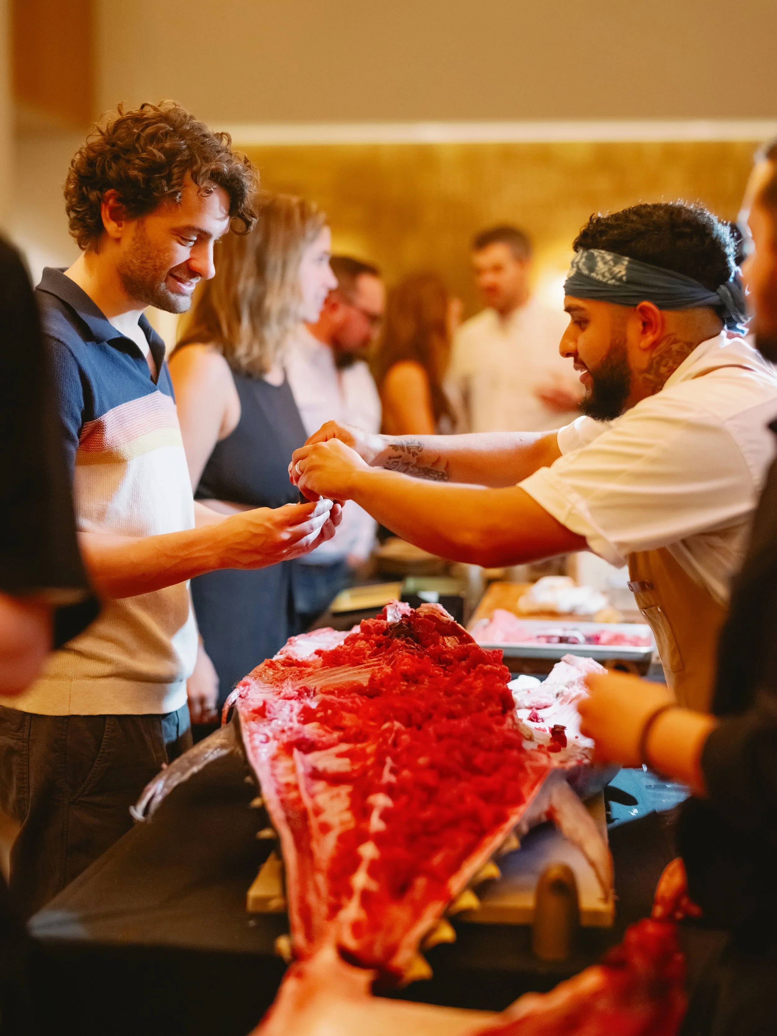 People at a social gathering selecting and sharing cuts of raw meat, including a large piece of beef, with a warm indoor setting.