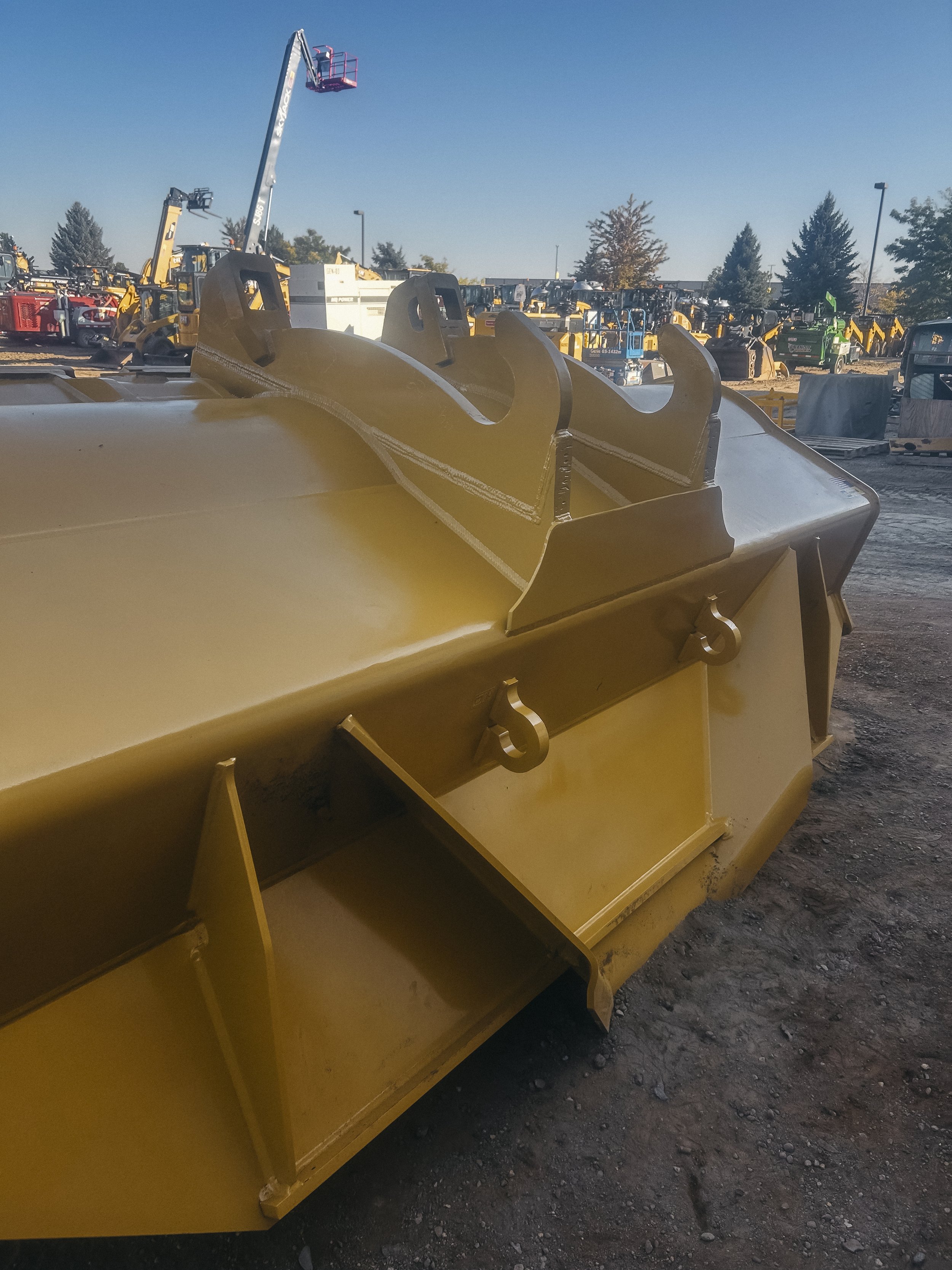 Close-up of yellow construction equipment attachment with hooks, on a dirt lot with various construction machinery in the background.
