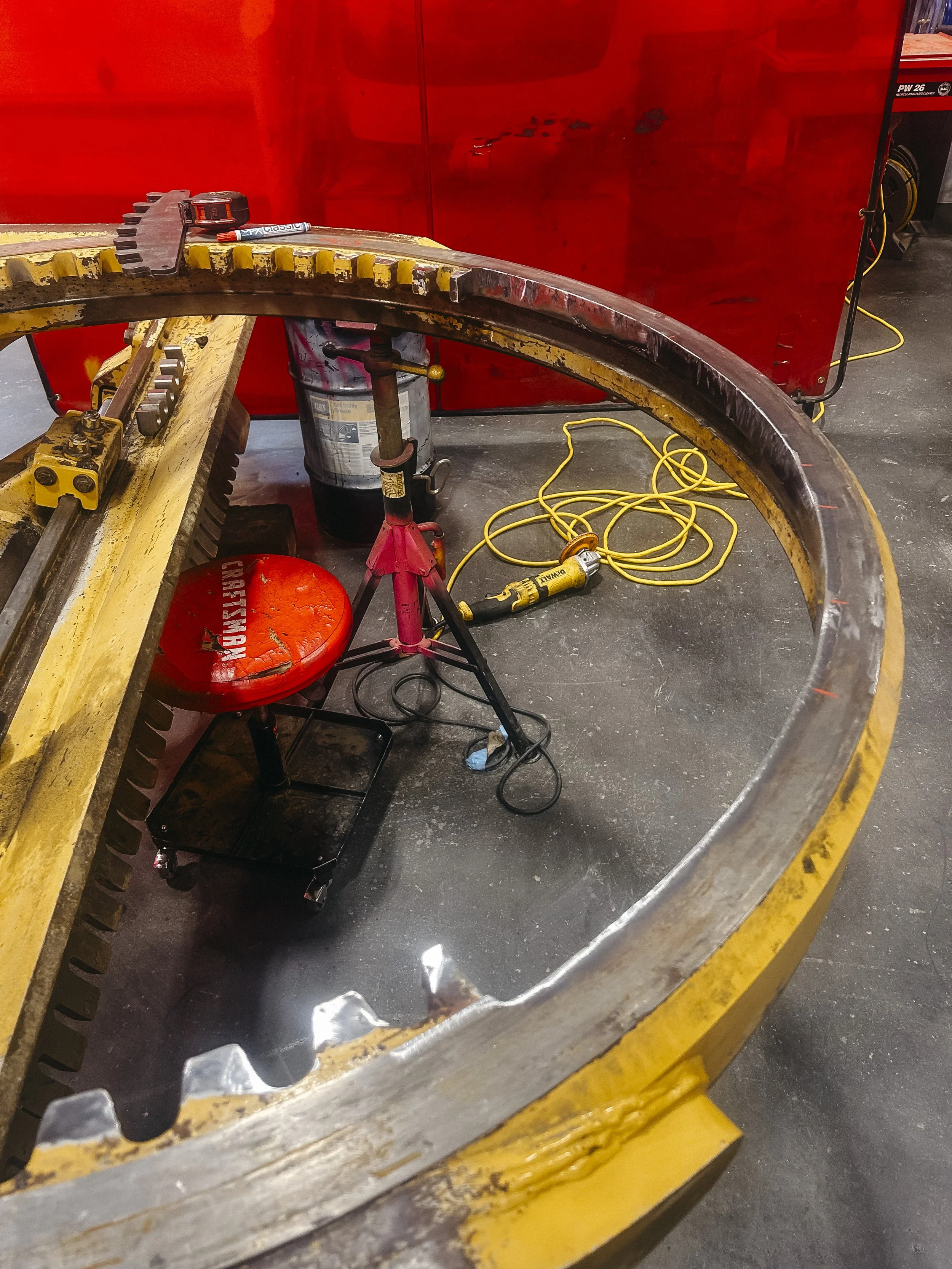 Large yellow gear with a broken edge on a metal worktable in a workshop, with tools, a red stool, and an air compressor in the background.