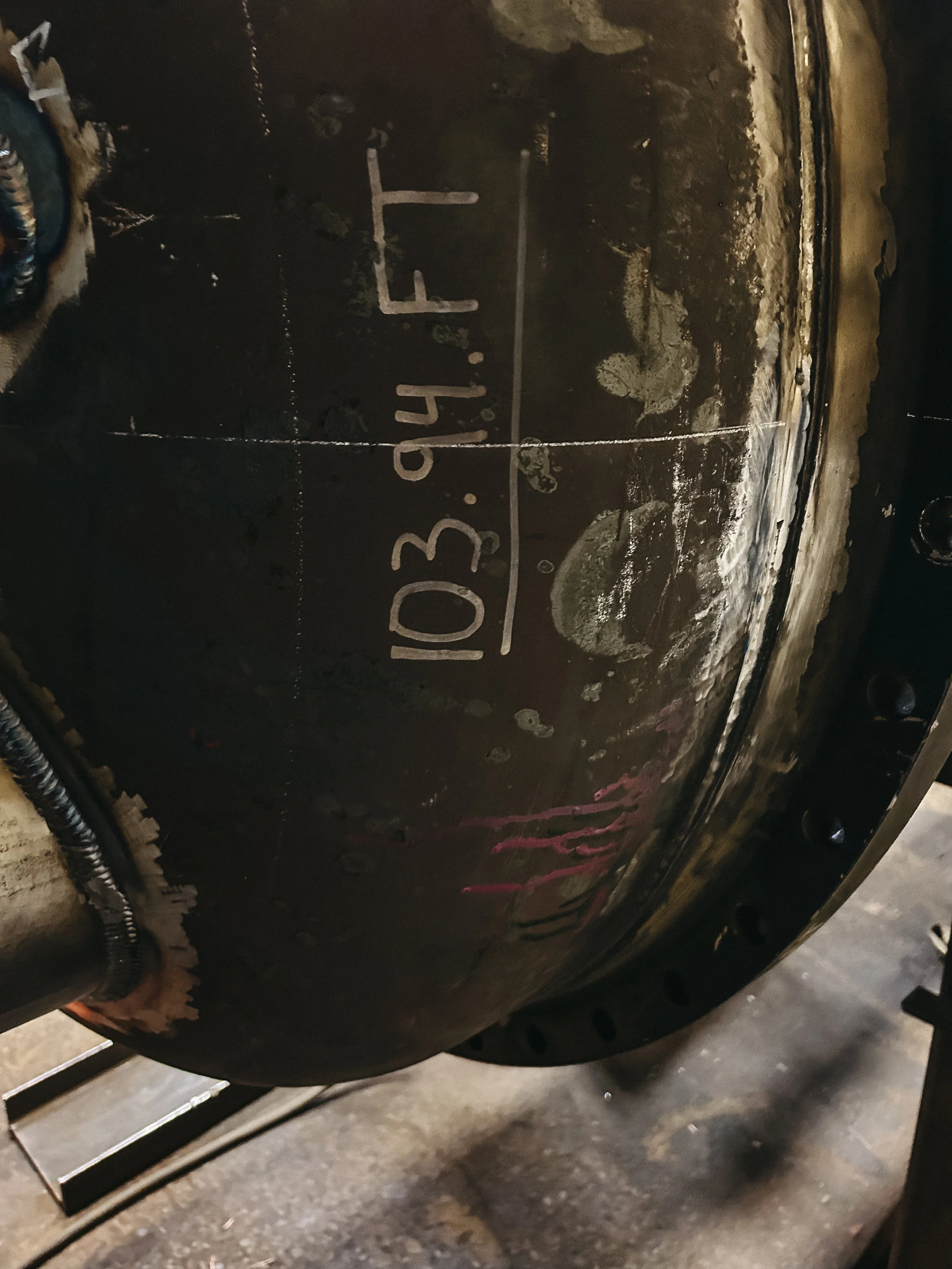 Close-up of a large metal tank or vessel with handwritten markings showing a date and some numbers, with some scratches, rust, and dirt on its surface.