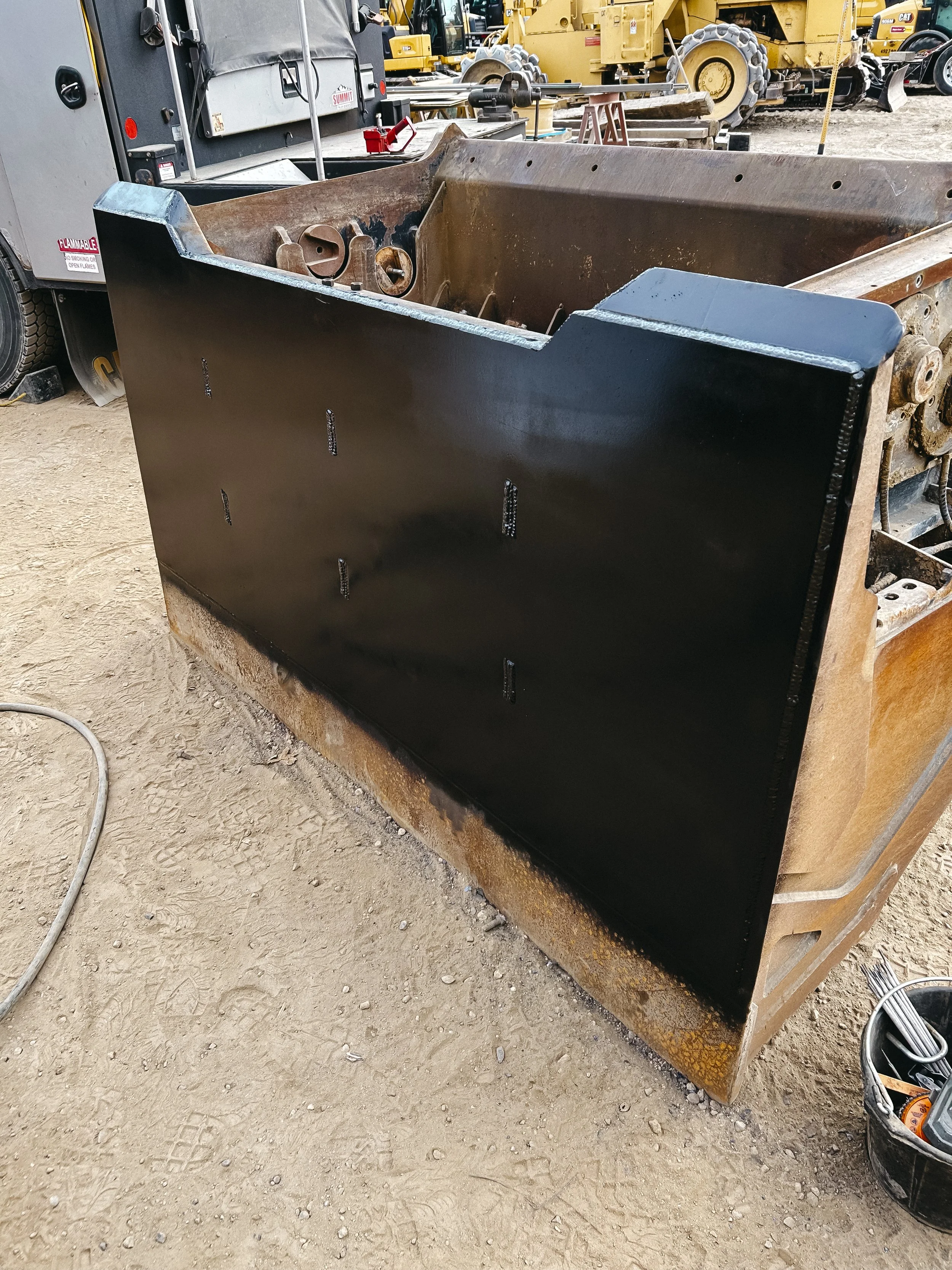 Close-up of a large, shiny black metal plate attached to a rusty construction machine at a construction site, with other construction equipment and dirt ground visible in the background.