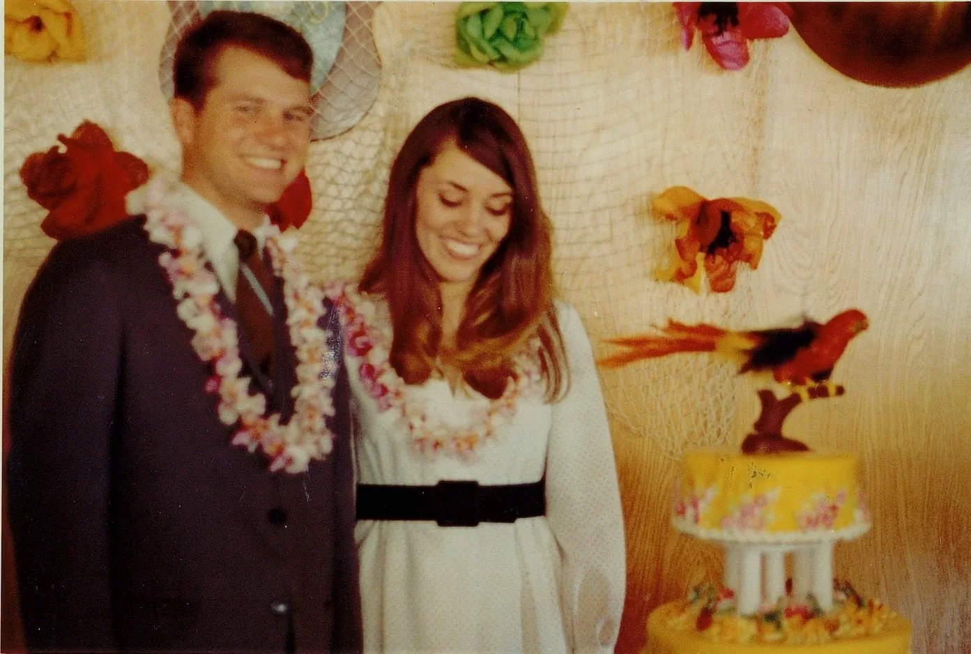 A man and woman are smiling at a celebration, wearing flower leis, with a decorated birthday cake featuring parrot toppers in front of them.