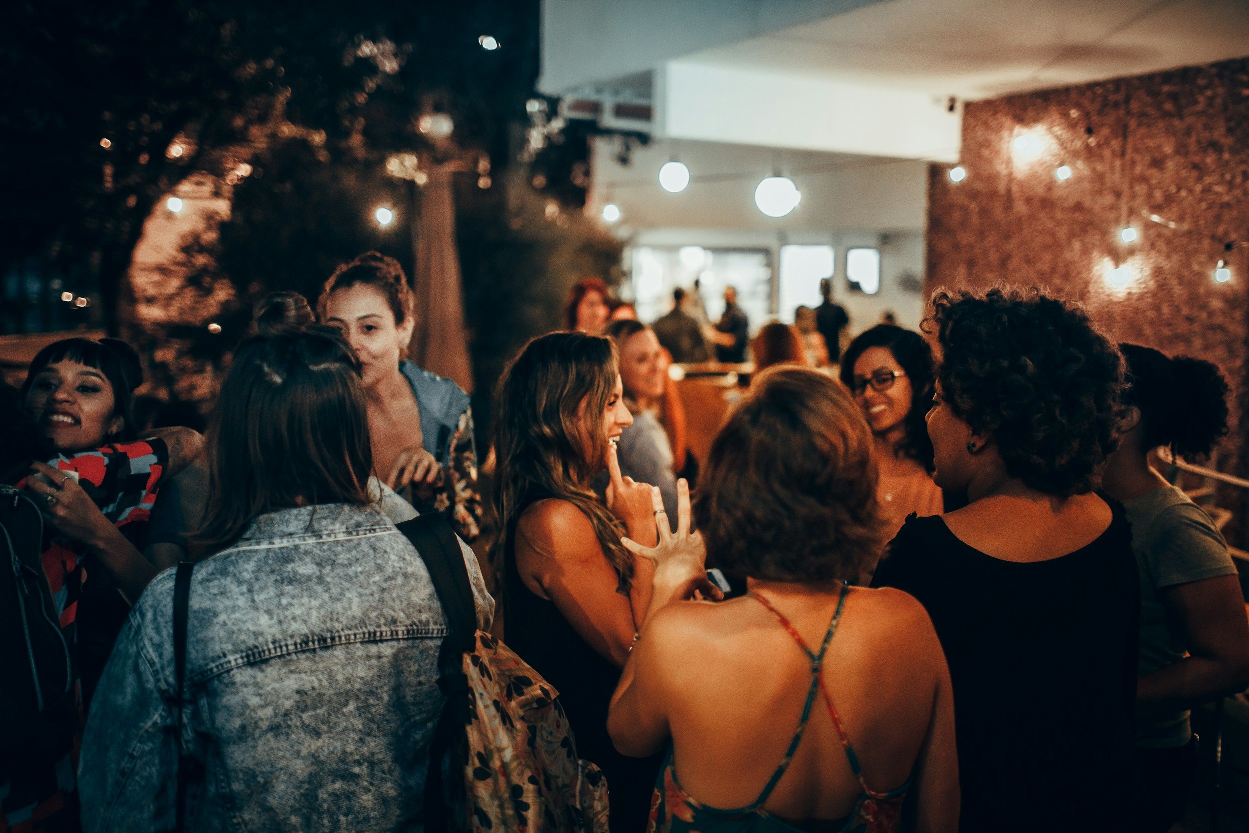 A group of diverse women socializing and dancing in a warmly lit indoor space at night. Some women are smiling and talking, holding drinks, with a background of hanging lights and a window.