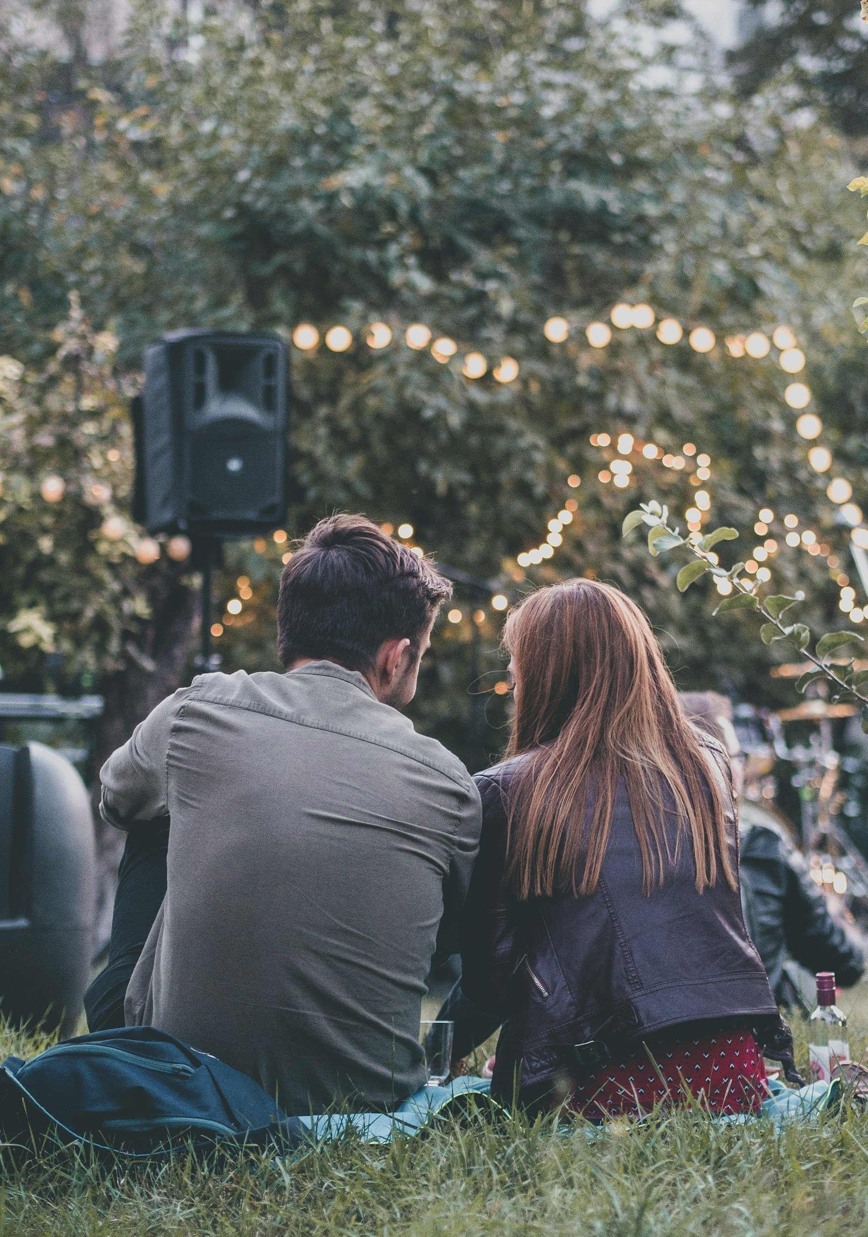 A couple sitting on the grass at an outdoor event with string lights hanging above, seen from behind. The scene is relaxed with trees in the background and a black speaker on the left.