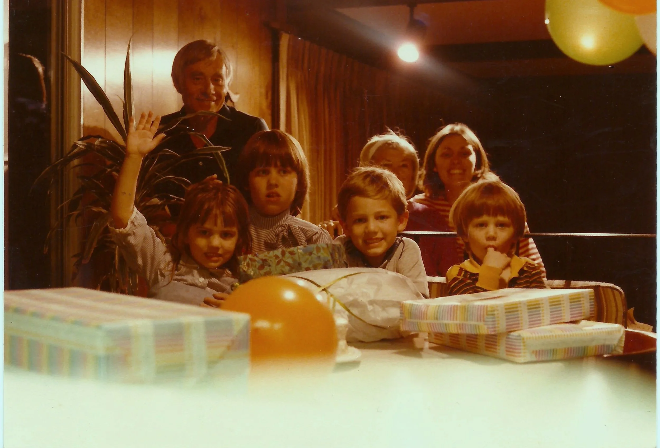 A group of children and adults at a birthday party with wrapped presents and balloons. The people are smiling, with some waving, in a room with wood-paneled walls and warm lighting.